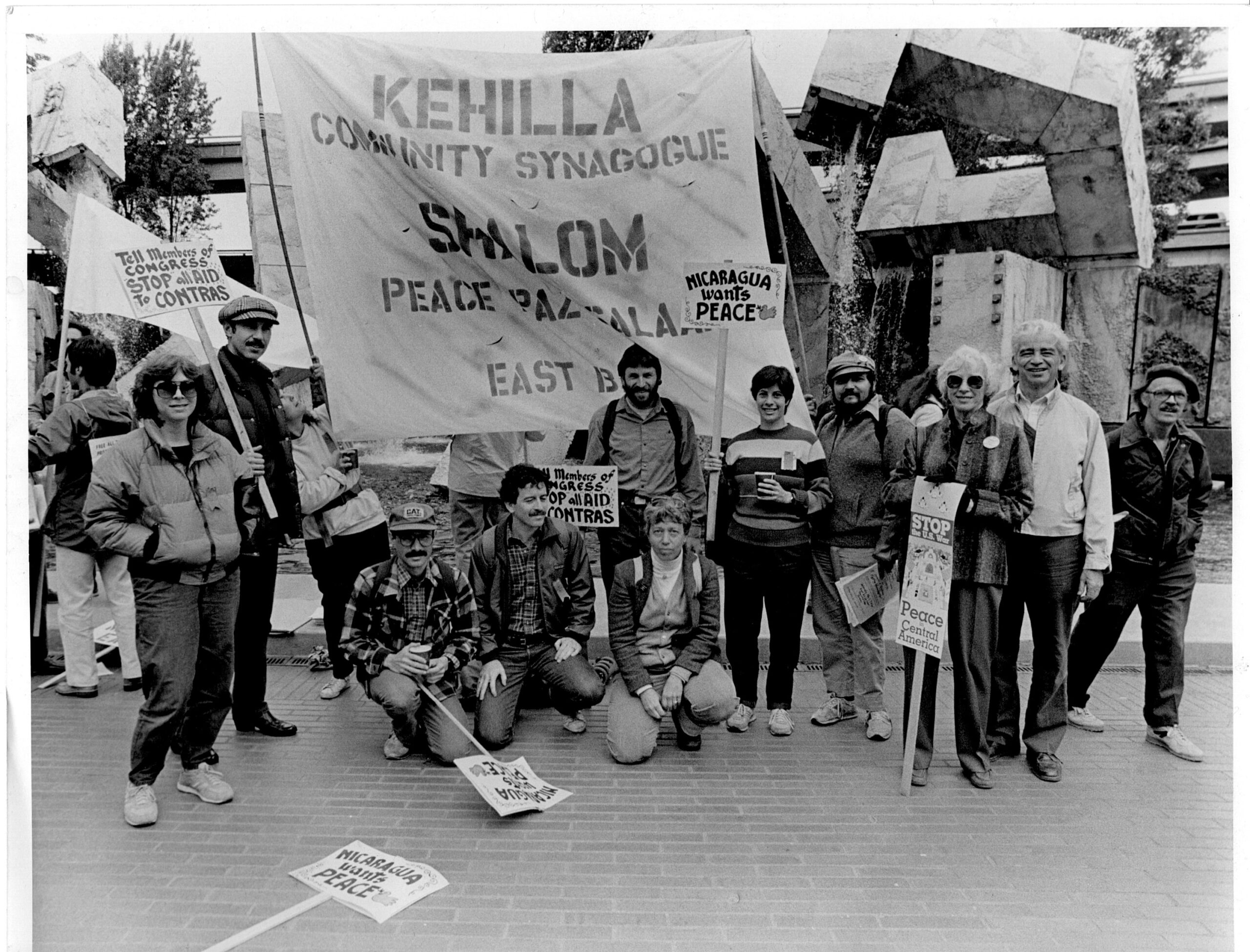 Protest signs say things like "Tell Members of Congress, Stop All Aid to Contras"