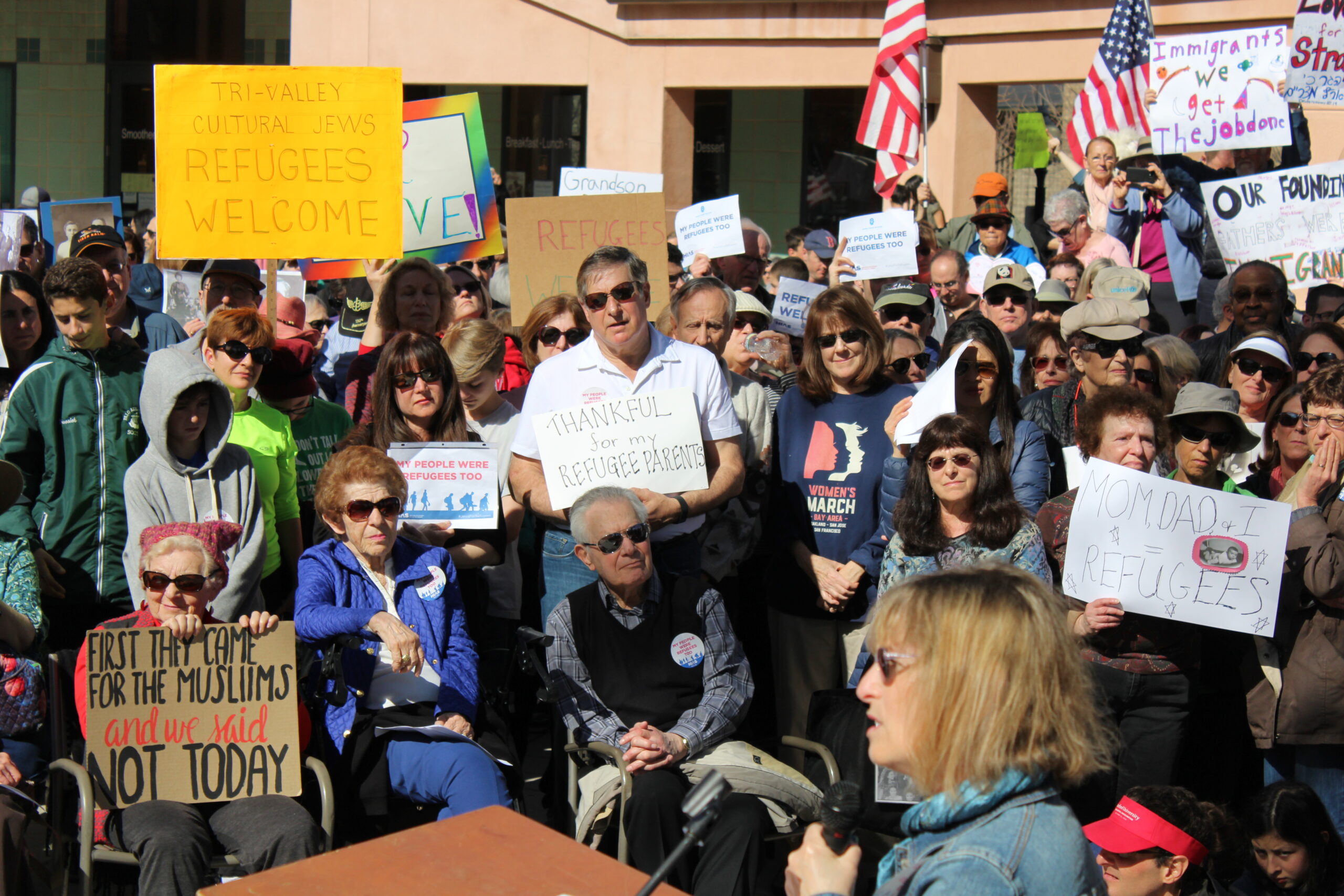 a large crowd holds signs like "tri-valley cultural jews: refugees welcome"