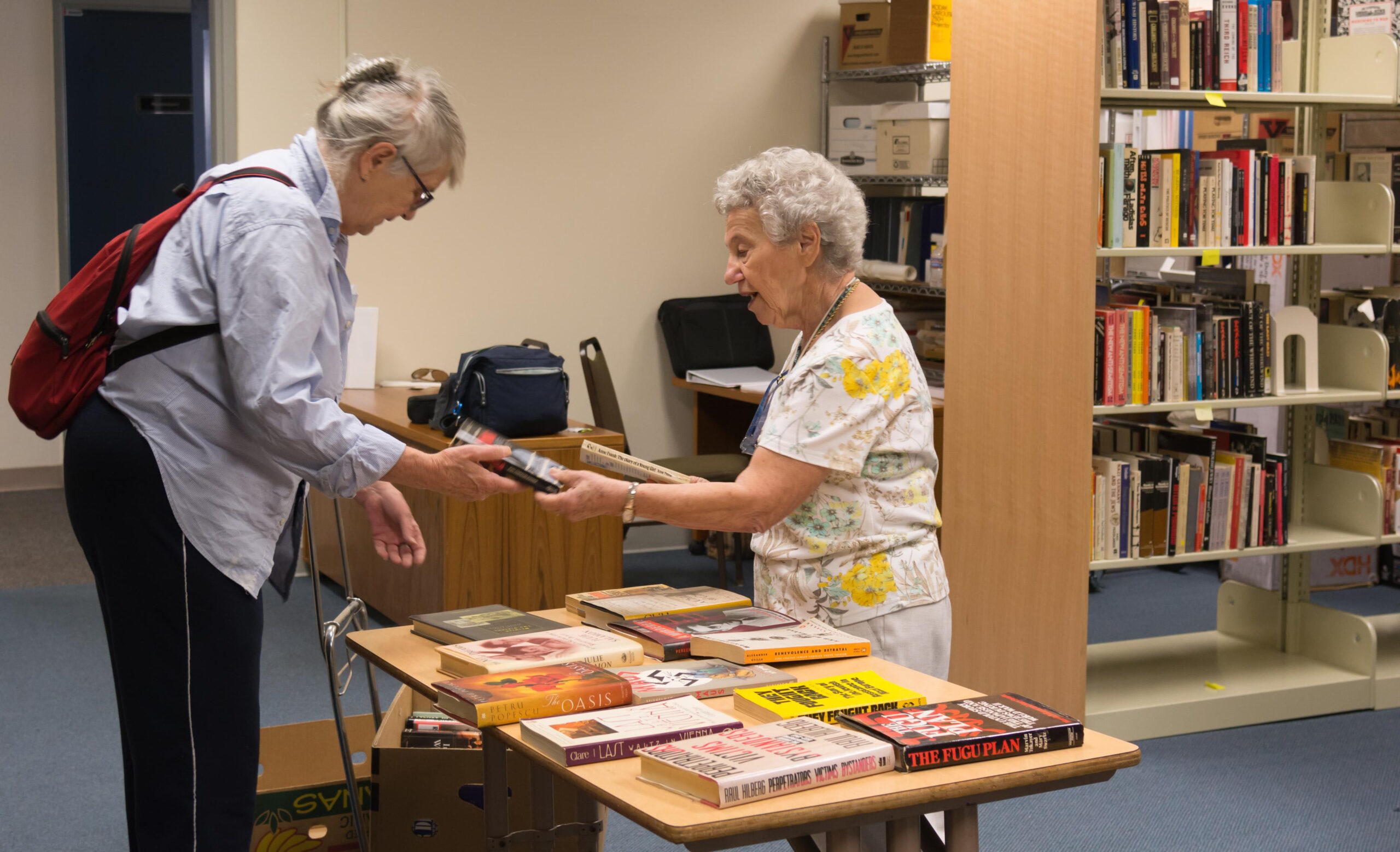 in a library, one old woman hands another woman a book