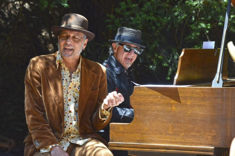 the two men sit at a piano together singing in the sunshine