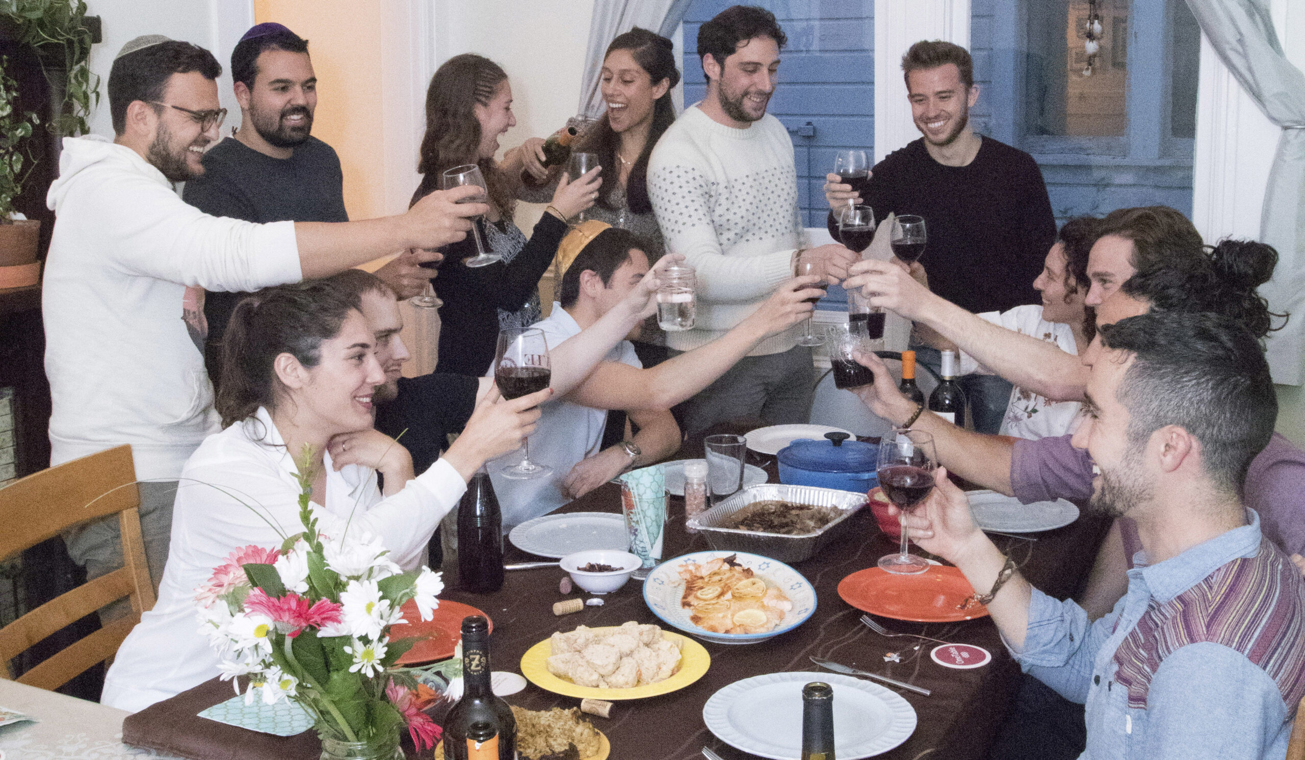 a dozen or so people stand and sit around a table raising glasses of wine
