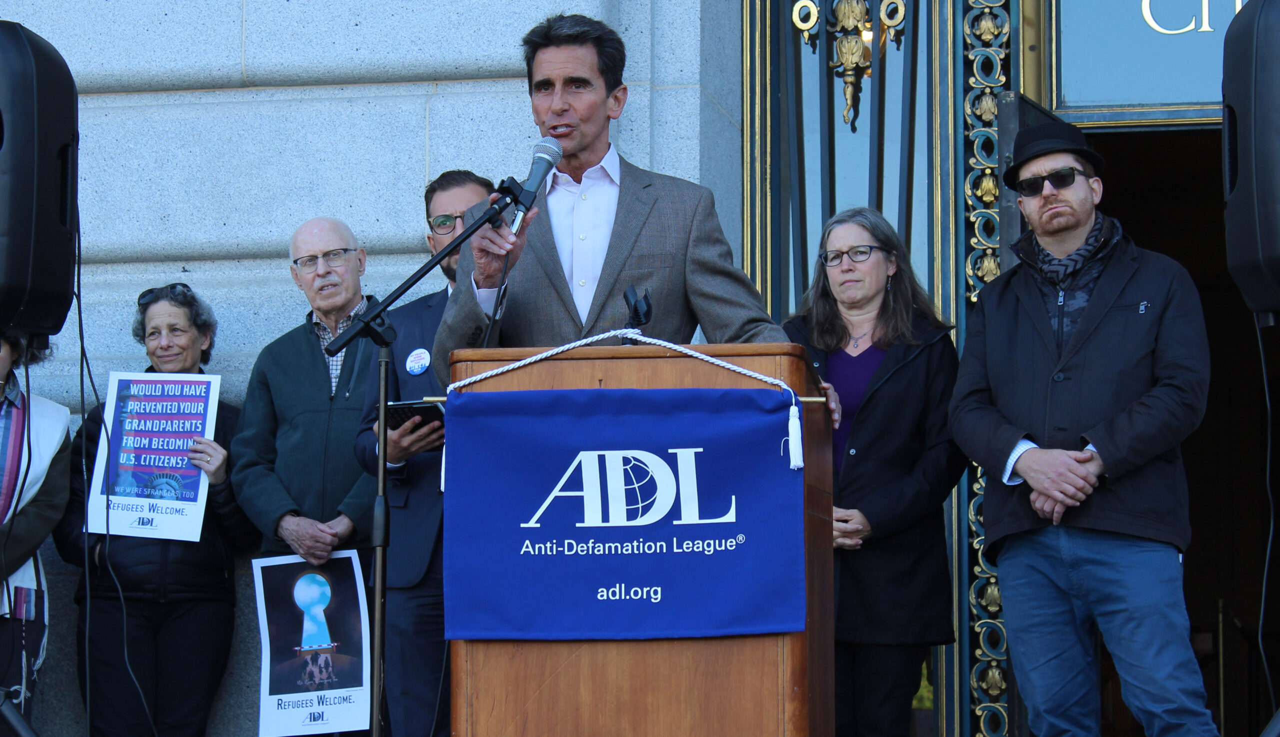 Leno speaks at a podium flanked by supporters with signs about welcoming refugees