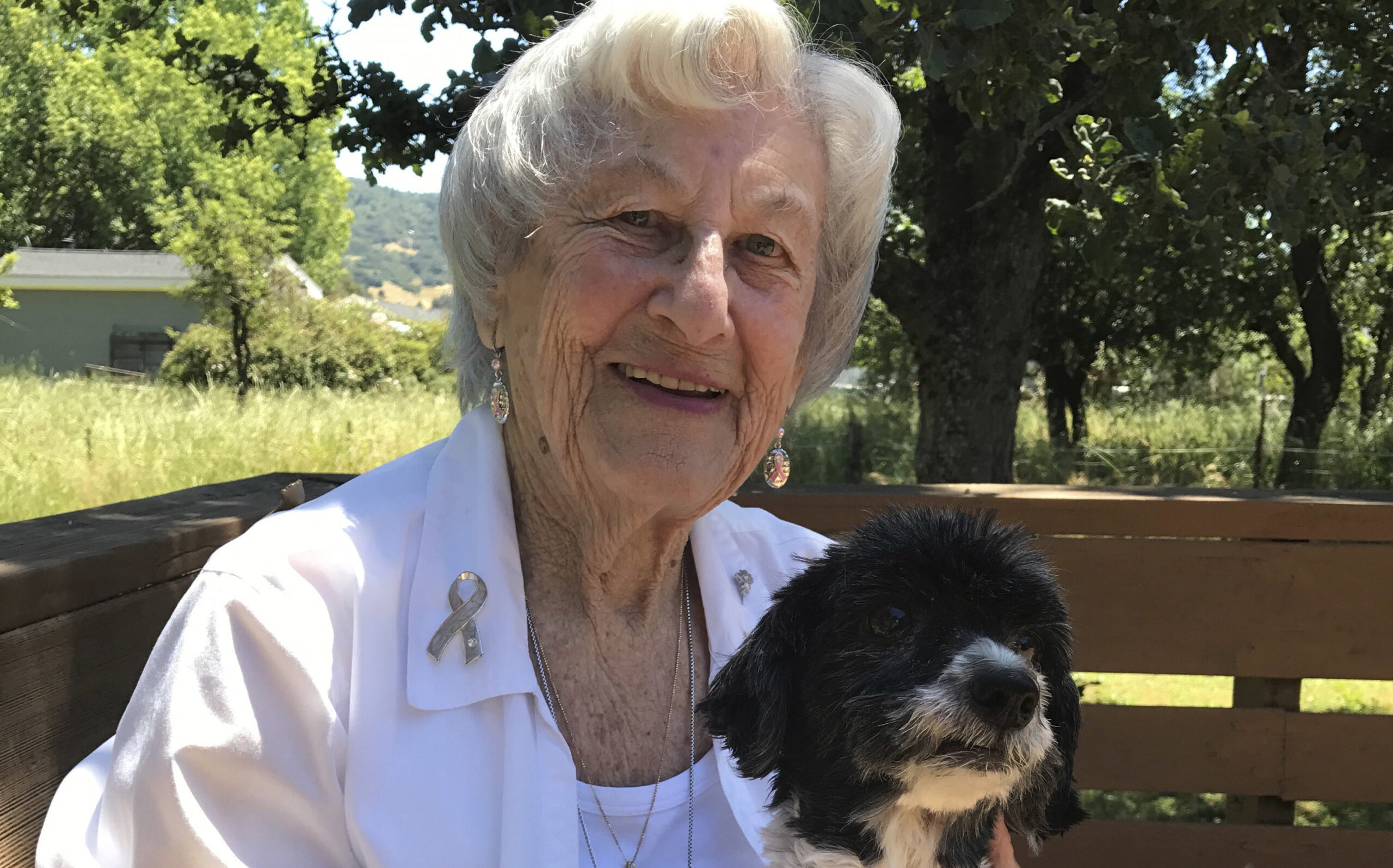 and old woman sits smiling on a park bench with a small black and white dog on her lap
