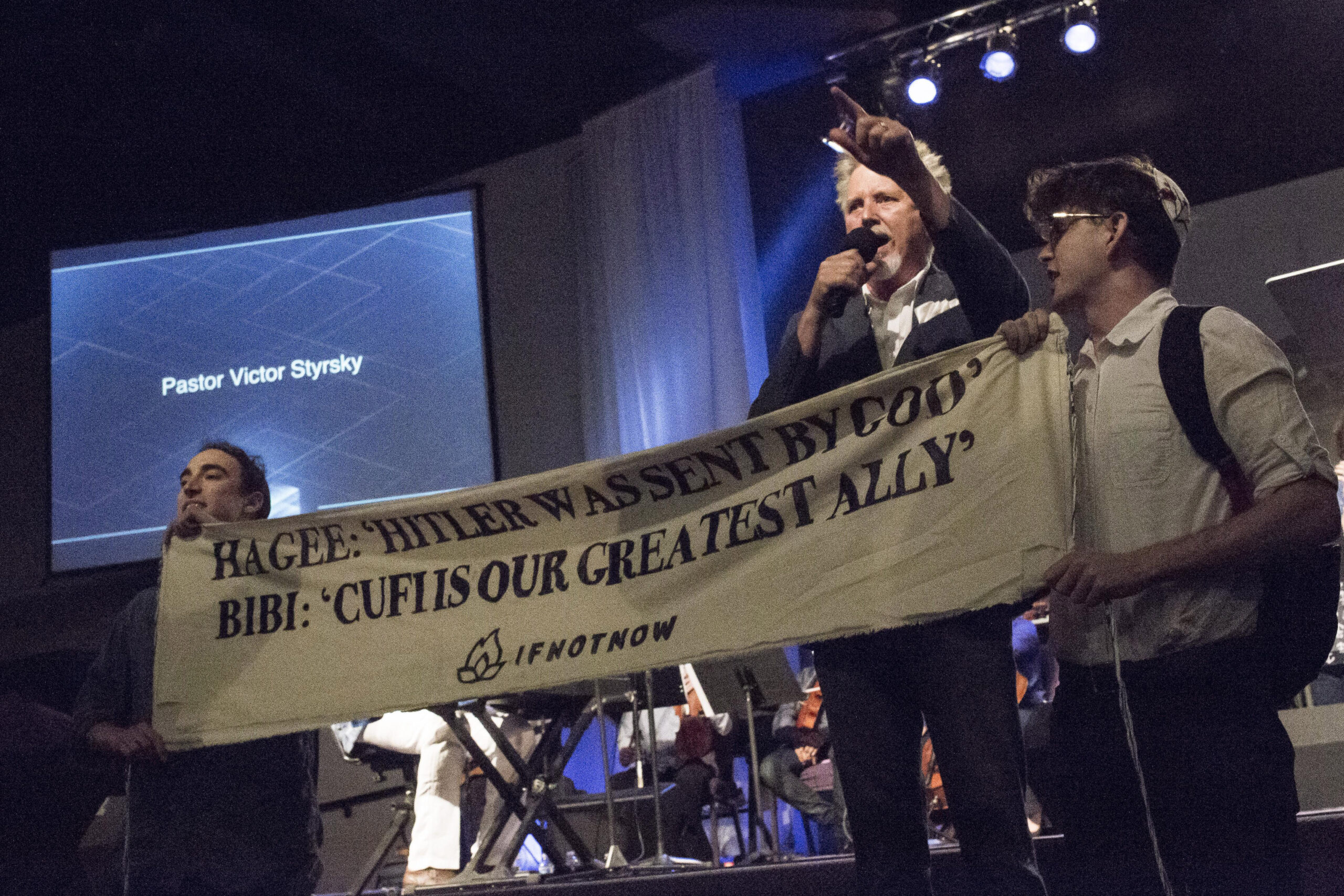 the protesters hold up a banner in front of the speaker. banner says "Hagee: 'Hitler was sent by God'" and "Bibi: 'CUFI is our greatest ally.'"