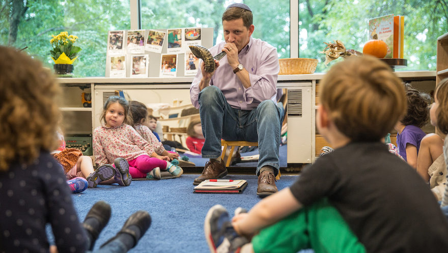 a man sits before preschool kids blowing a shofar