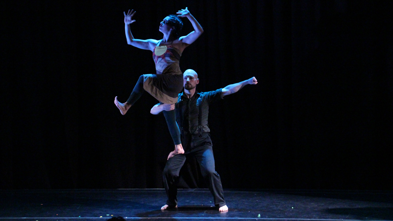 in dramatic blue lighting, an acrobatic couple do an improbable thing with each other's bodies
