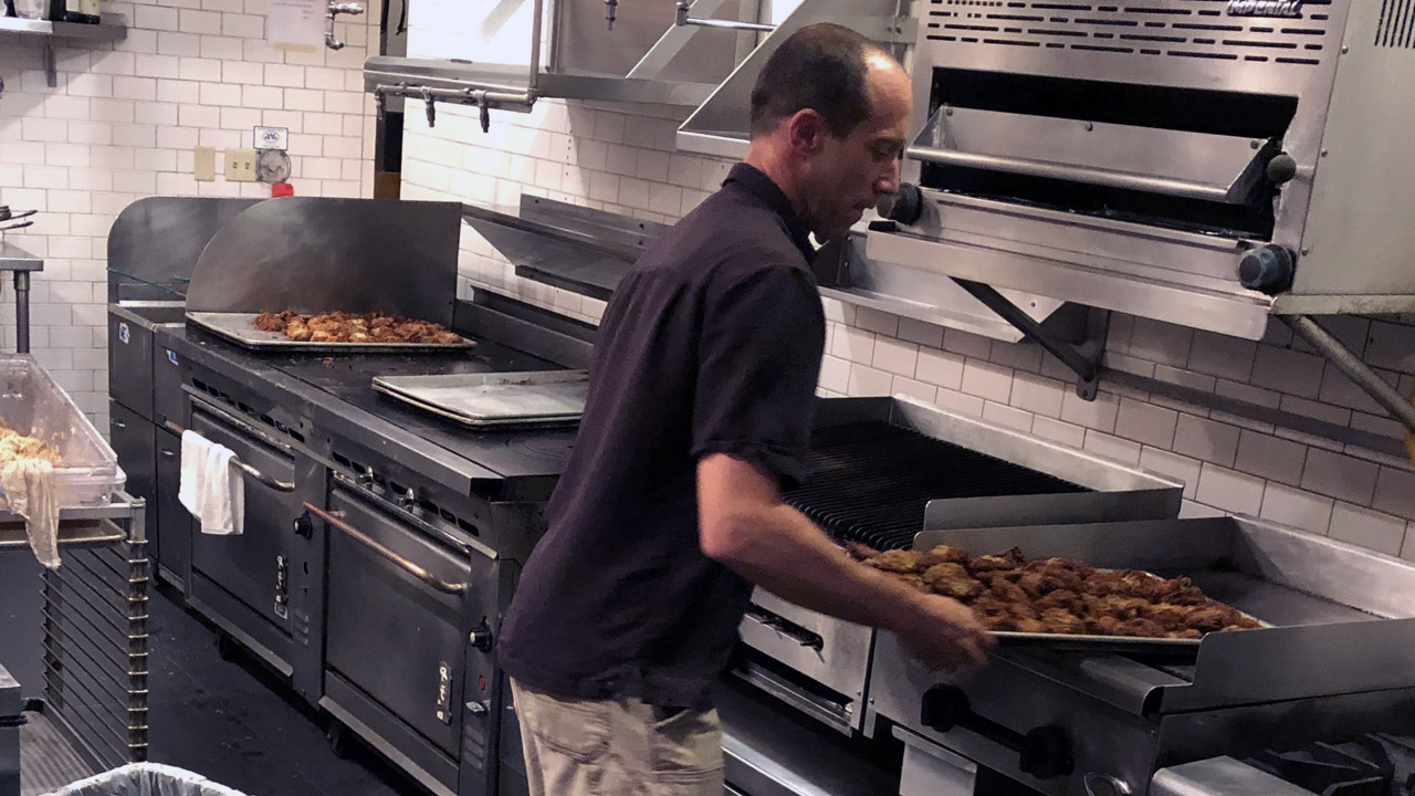a man in a commercial kitchen holding a large tray of latkes
