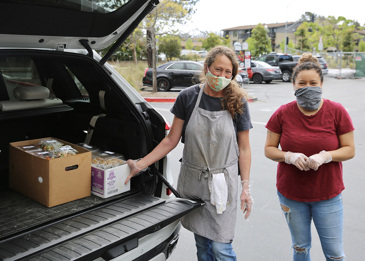 Two women with face masks on stand at the back of an SUV with it's rear door open and boxes of food inside