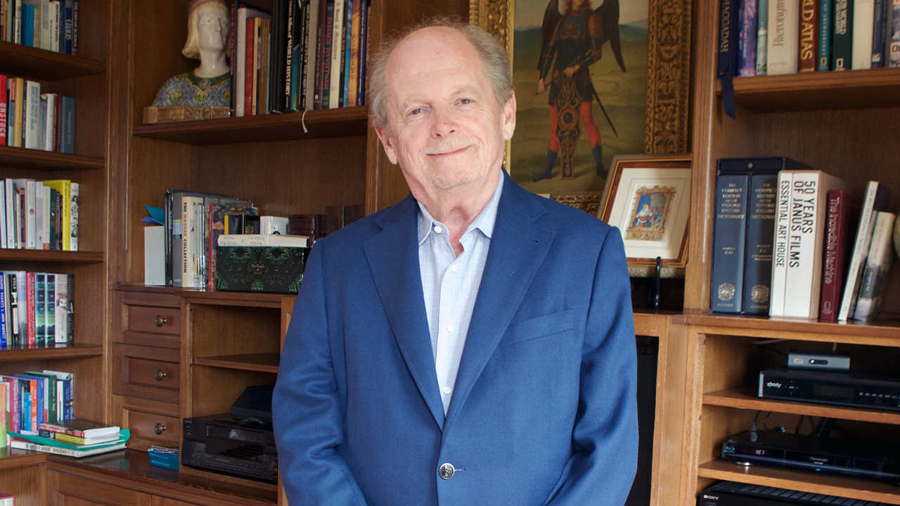 an old bald man stands in a well-appointed book-lined study