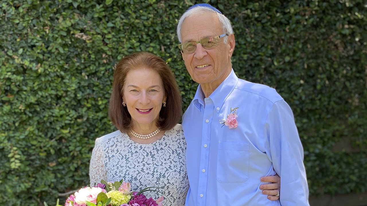 an older white couple standing together, smiling. she is wearing a white dress and holding a bouquet. he has a blue shirt with a flower pinned to it.