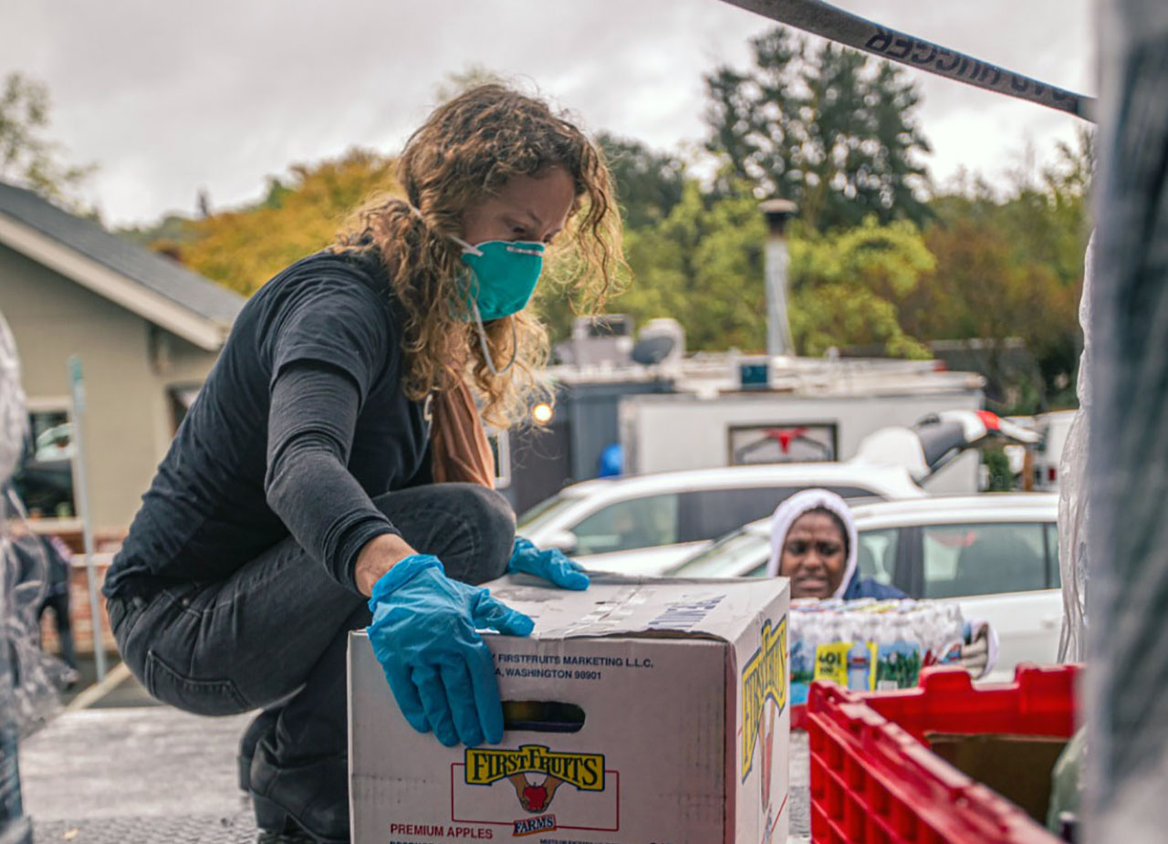 A woman kneels in the back of a large delivery truck to pick up a box of produce