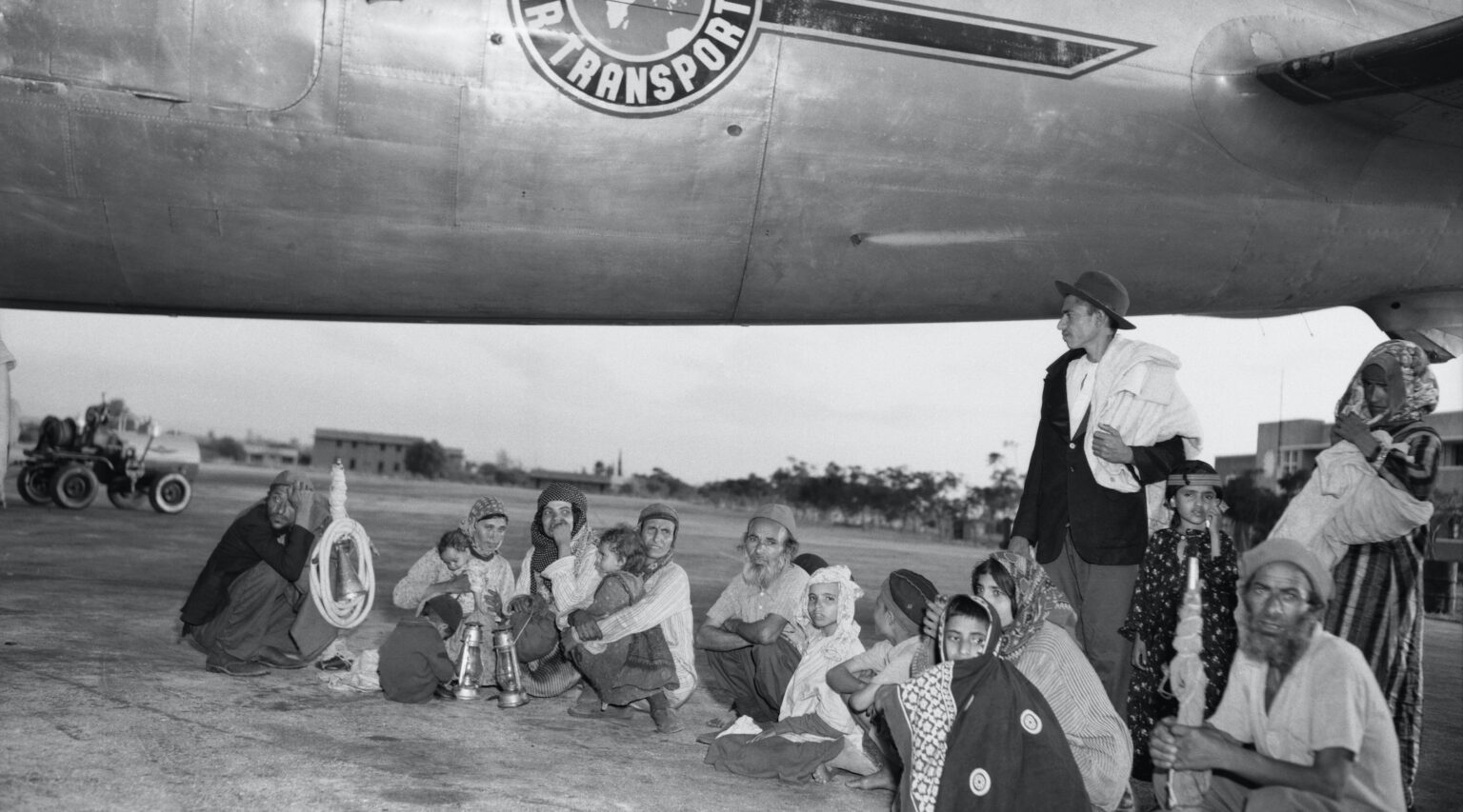 Black and white: A group of men, women and children sit on the tarmac near an old airplane