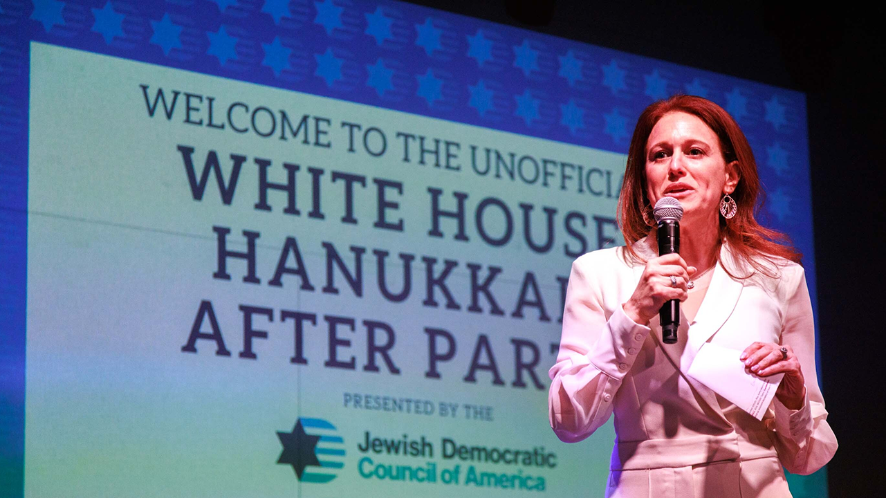 picture of Halie Soifer standing on stage with a sign behind her that says "welcome to the unofficial white house hanukkah after party"