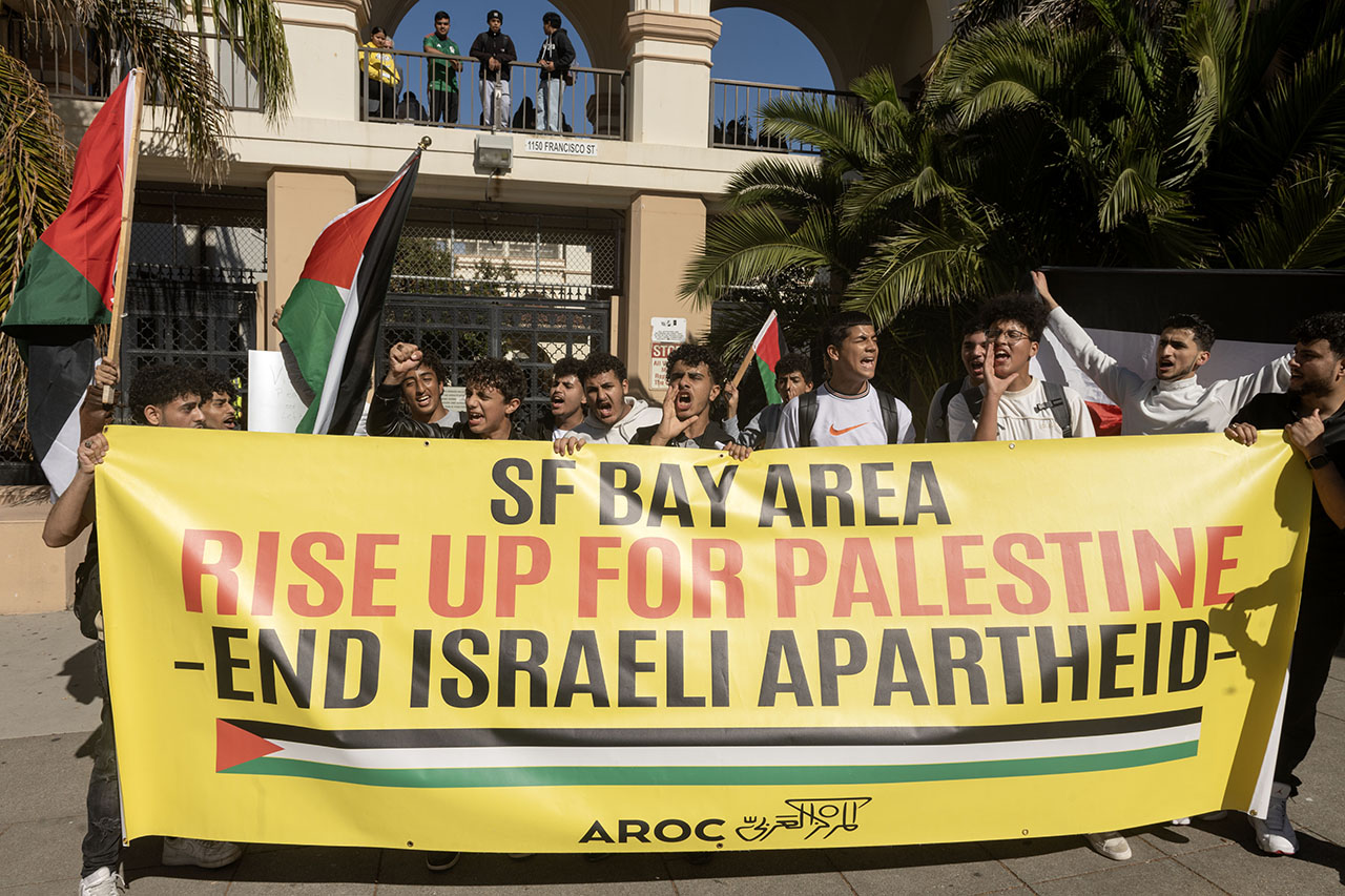 A group of protesters. Some hold up Palestinian flags. Others are holding up a large yellow banner that says "SF BAY AREA, RISE UP FOR PALESTINE, END ISRAELI APARTHEID"