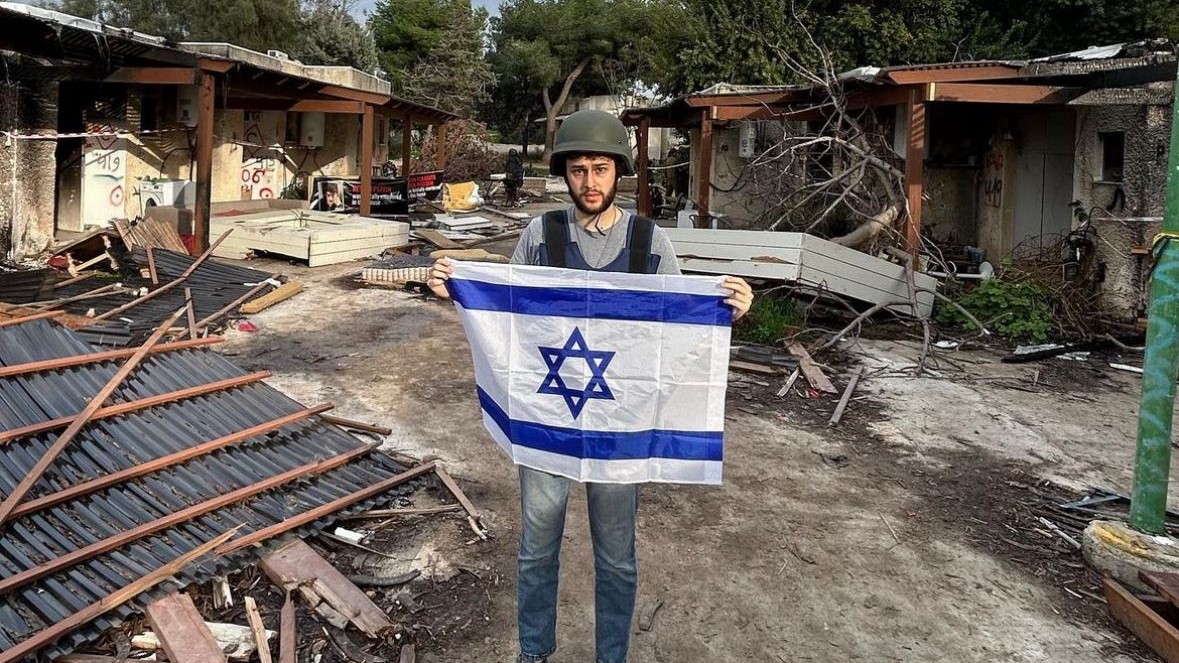 Man wears helmet and holds Israeli flag with destruction behind him
