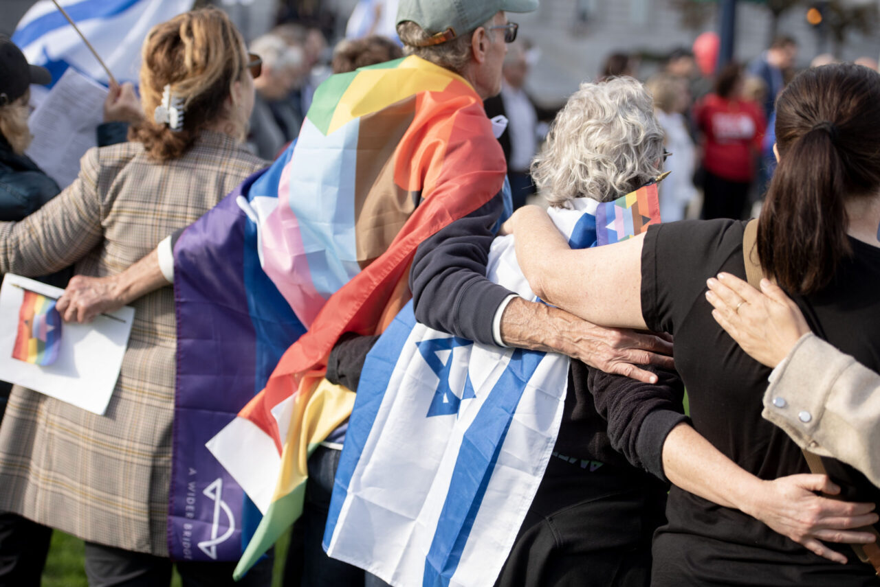 Backs of people standing with their arms around each other with Israeli and gay pride flags