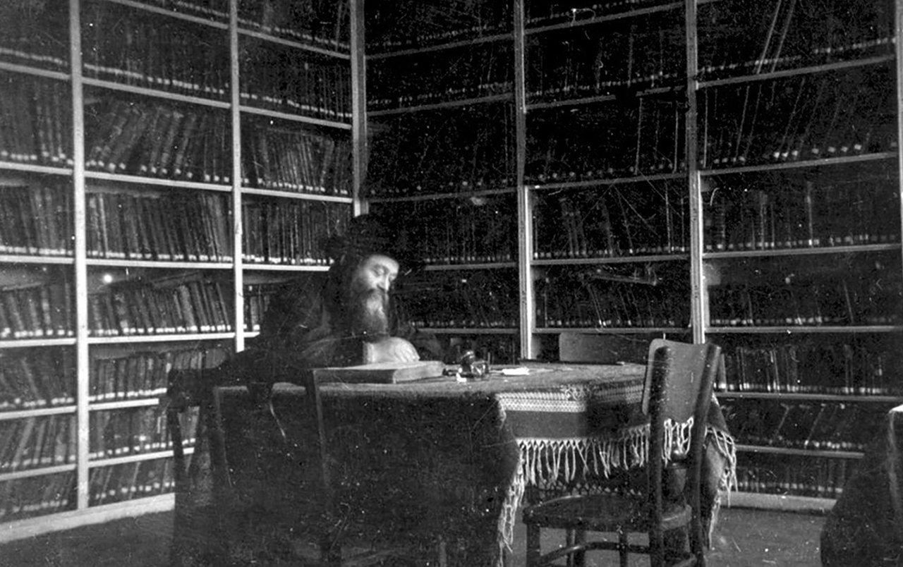an bearded man sits at a desk in a room lined with books