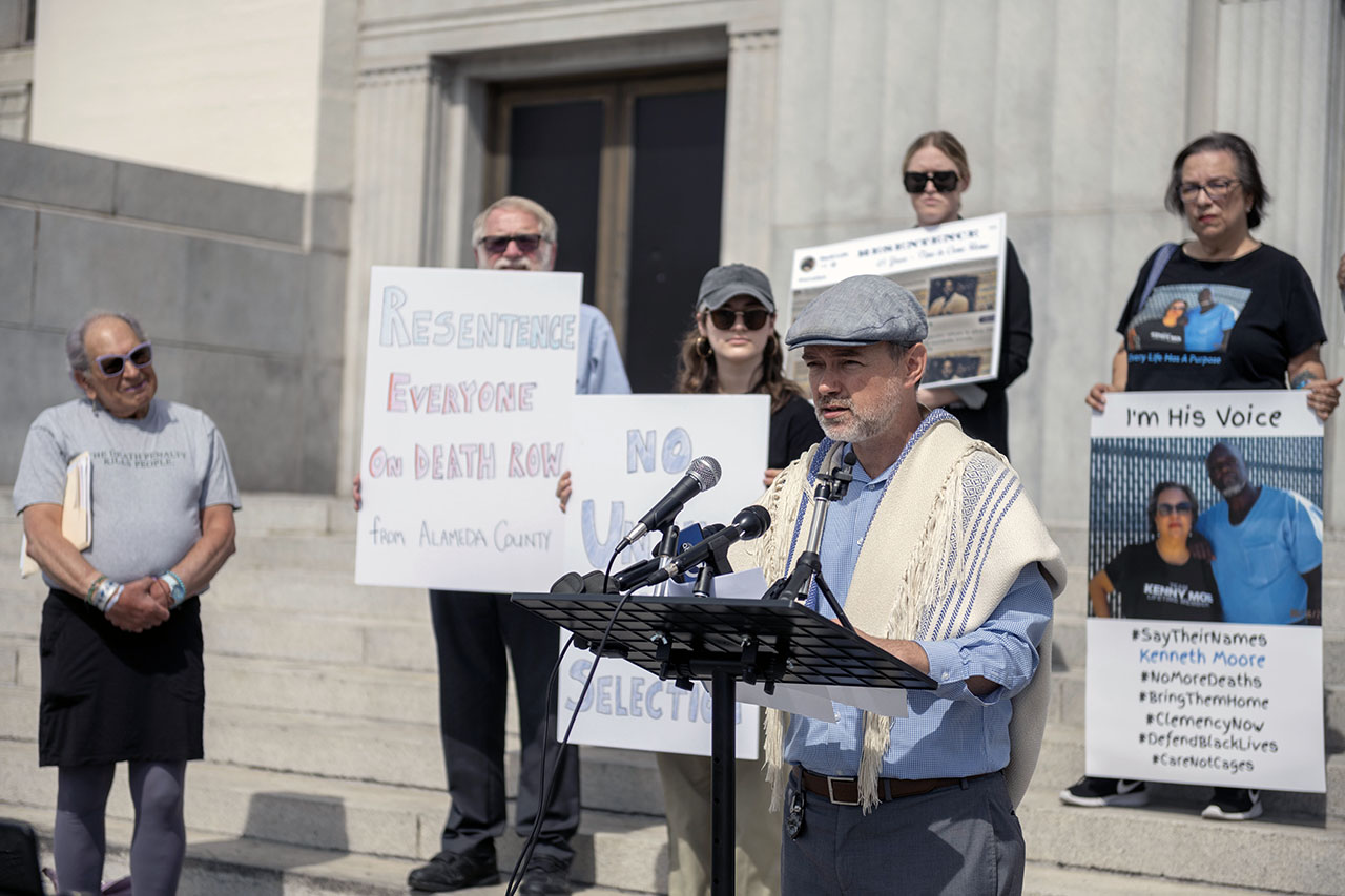 A white man wearing a tallit stands speaking at a podium on the courthouse steps