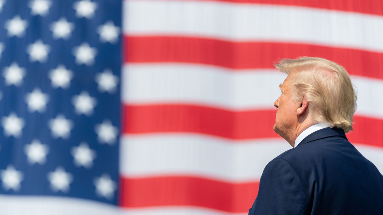 Trump seen from behind, standing before a giant American flag