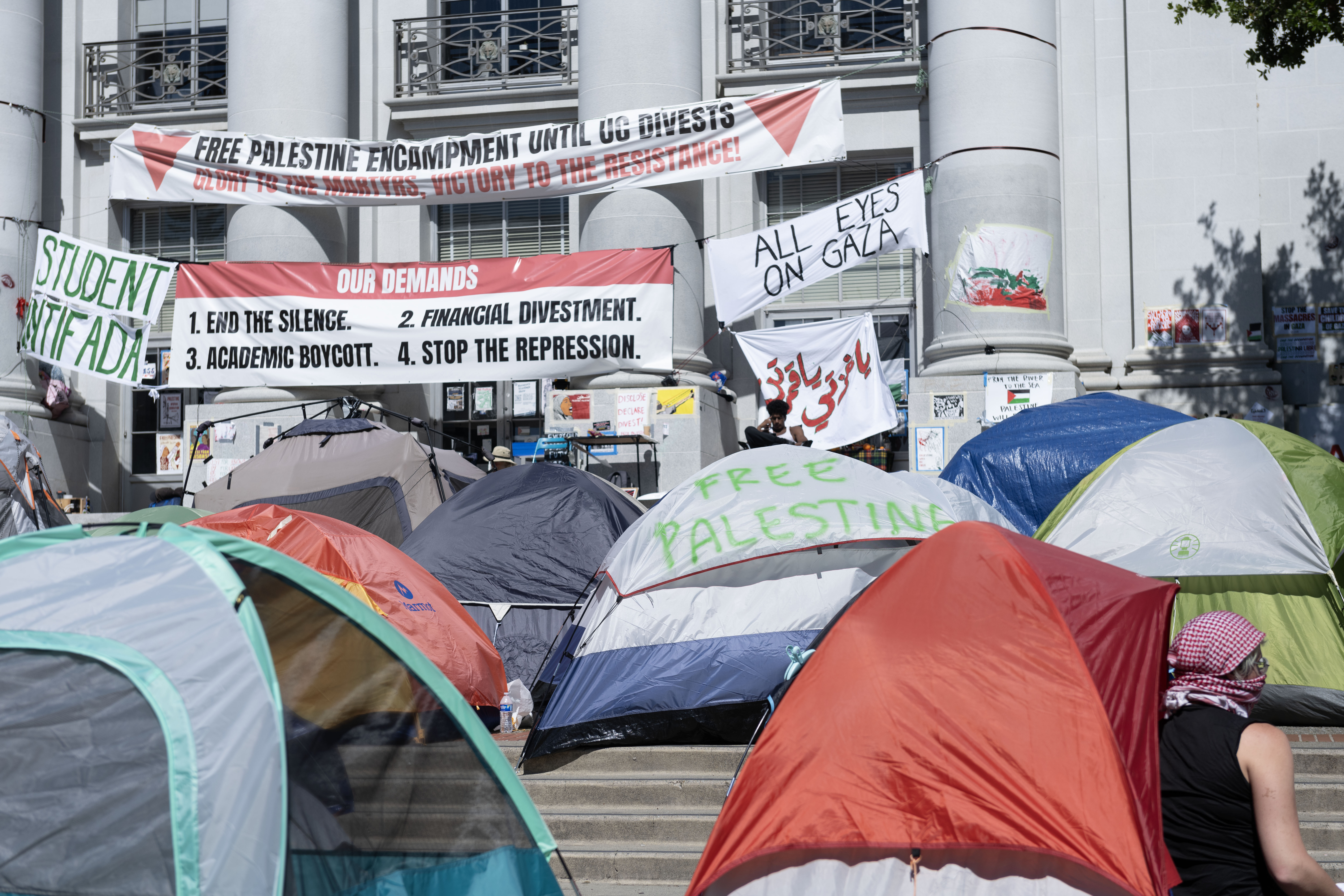 The steps of a large academic building are crowded with small tents and banners with messages such as "Glory to the Martyrs" and "Student Intifada"