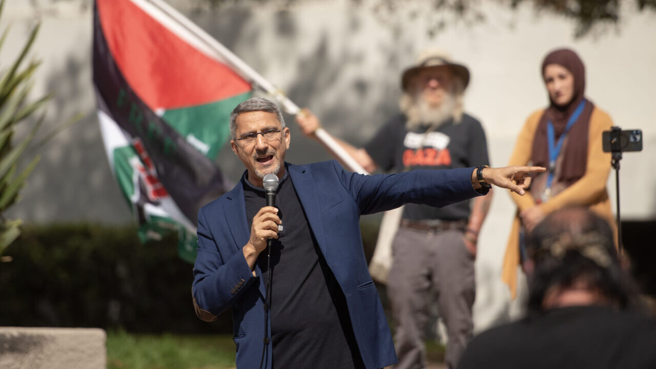 A man in a blue blazer holds a microphone as another man waves a Palestinian flag behind him