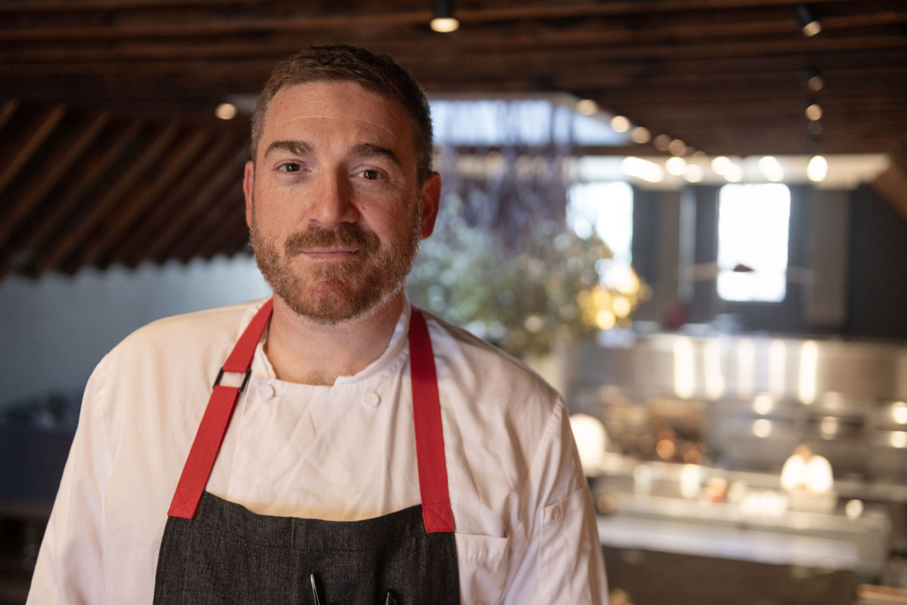 a smirking man with a short beard wears an apron - a restaurant dining room can be seen in the background