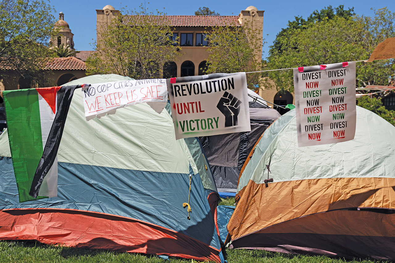Sings hanging from a line in front of several tents say "Divest Now" and "Revolution Until Victory"