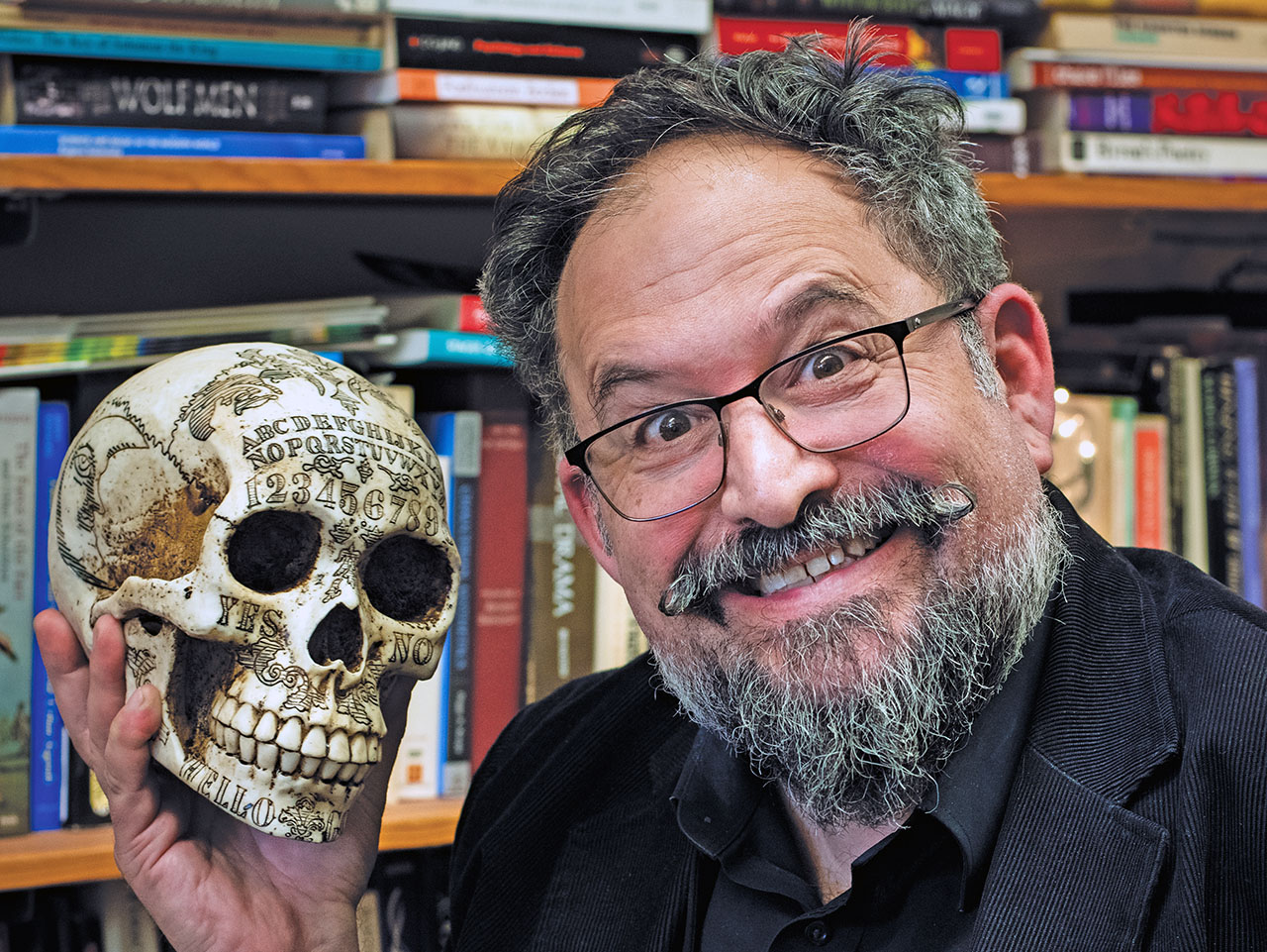 a man with a mustache and beard grins menacingly as he holds up a skull inscribed all over with letters and numbers