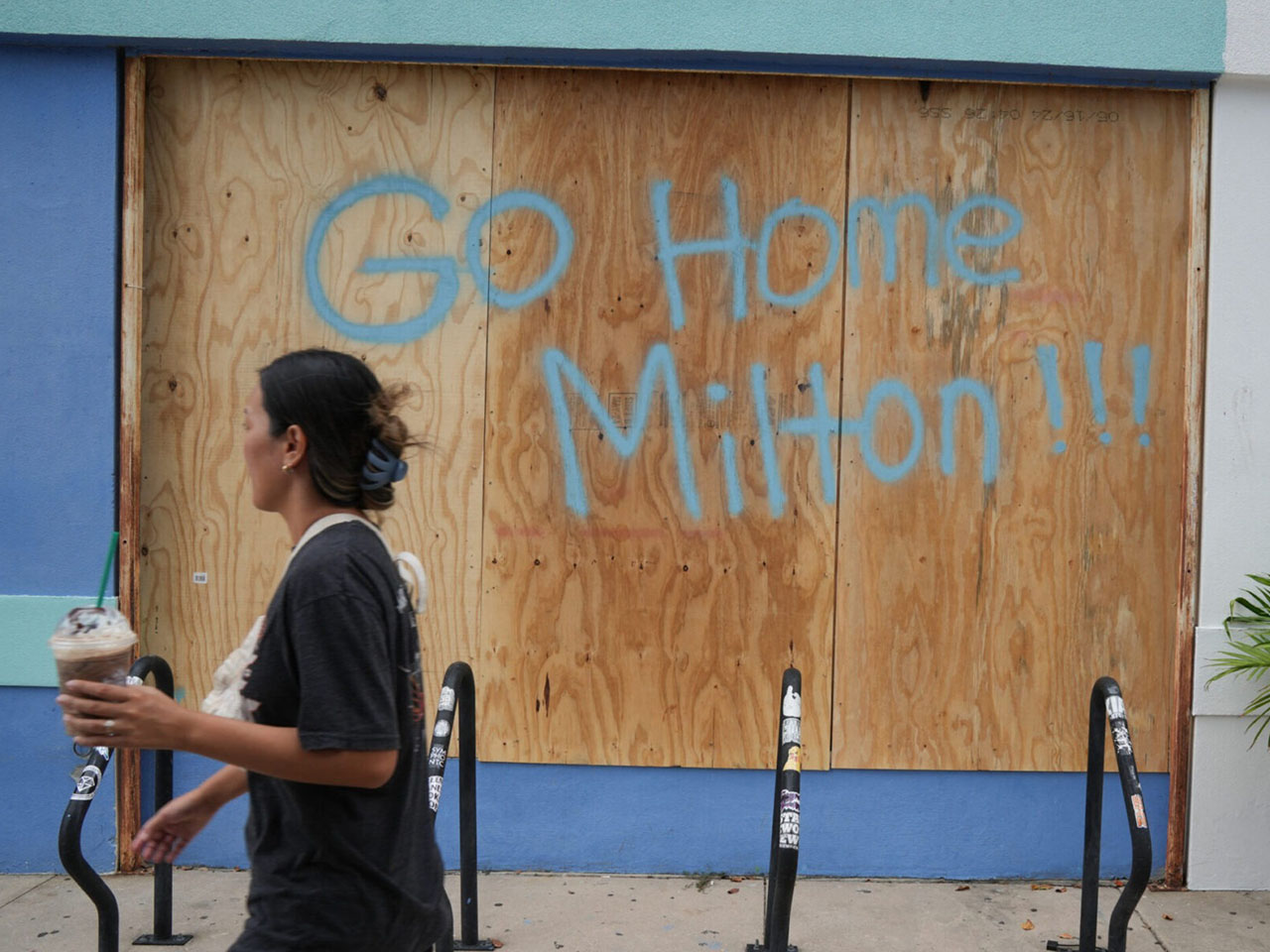 a woman walks past a boarded up storefront with "Go Home Milton!!" written on it