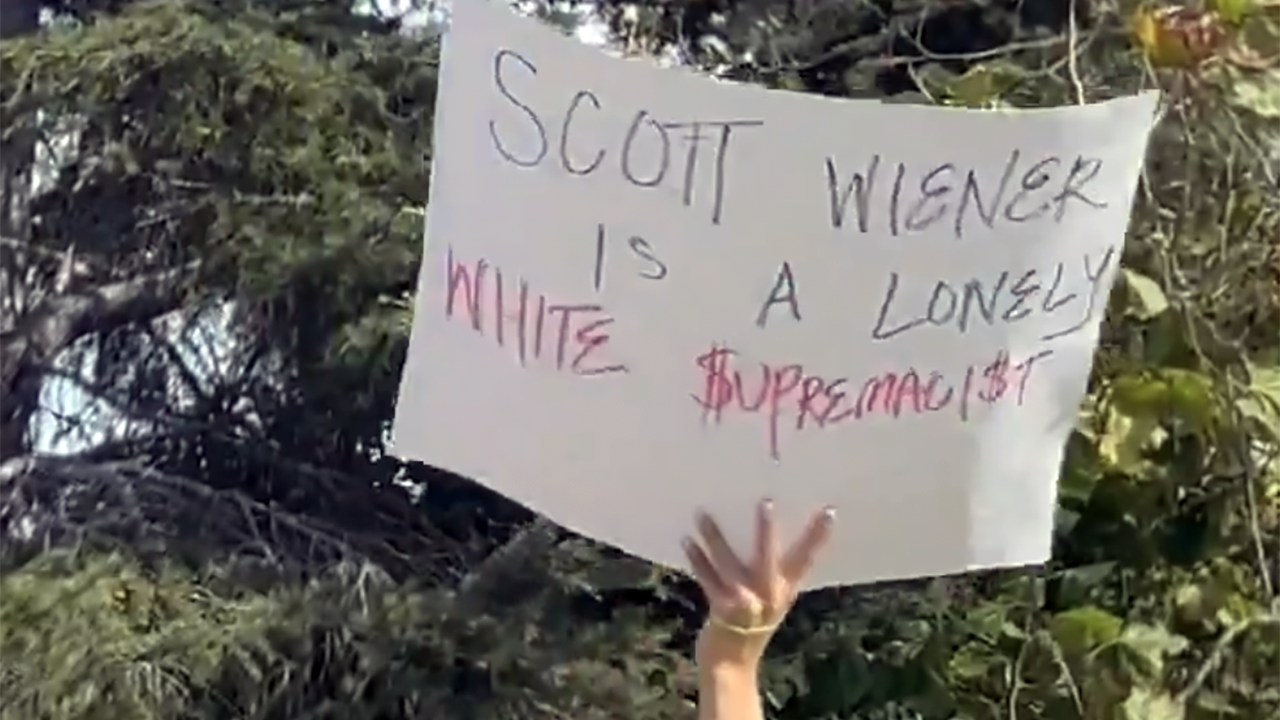 Outside a pumpkin carving event held by State Sen. Scott Wiener, a protester holds up a sign that reads "Scott Wiener is a lonely white supremacist." (Screenshot via @SFJCRC on X)