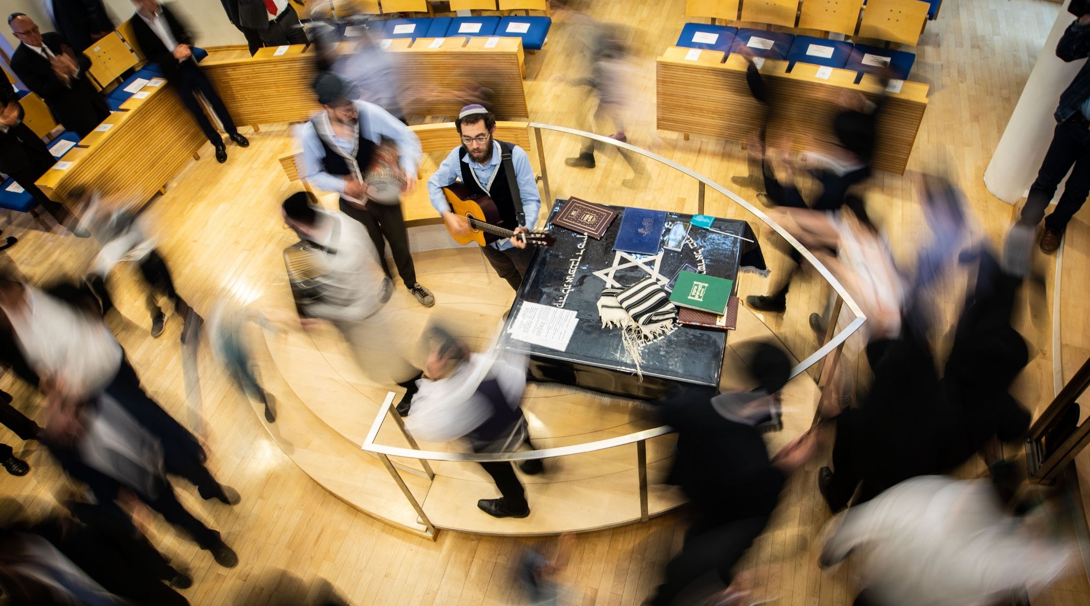 a timelapse photo shows blurs of people circling a bimah carrying Torahs as a guitar player stands in the center