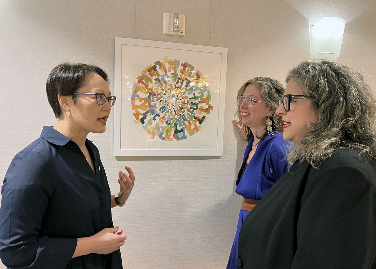 Three women stand talking near a circular piece of art made of Hebrew letters