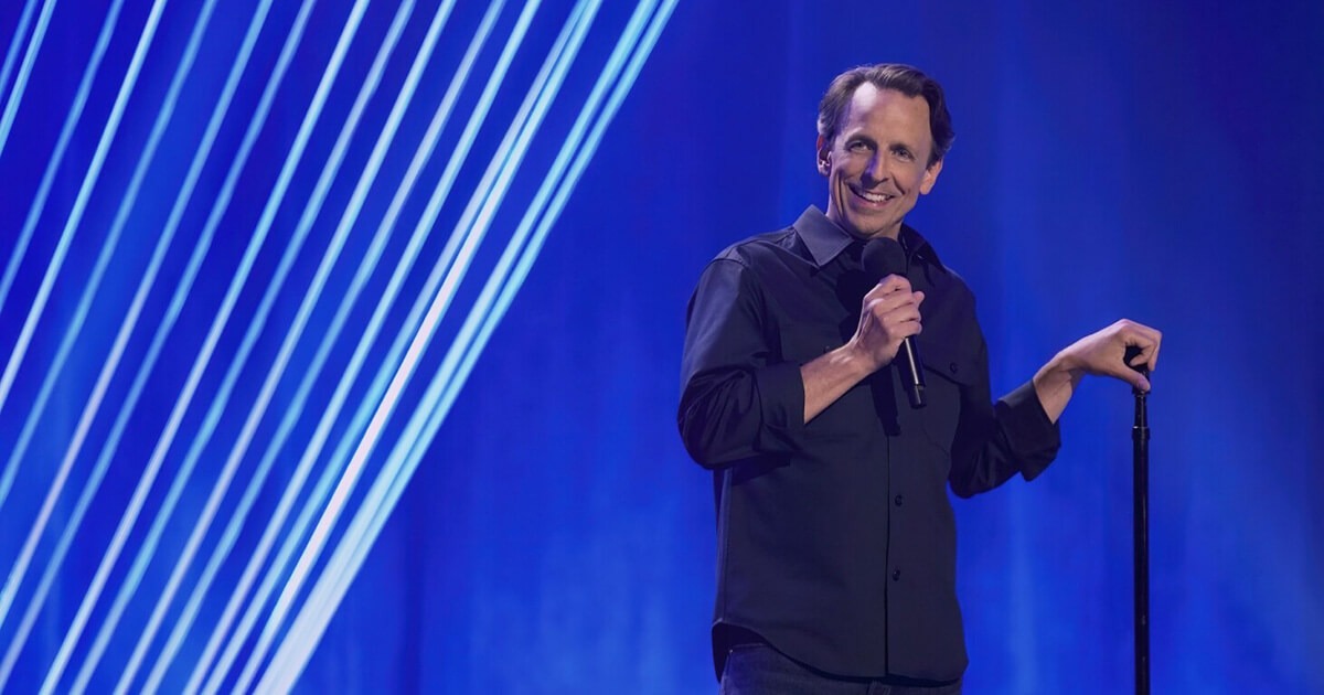 Meyers stands on stage holding a microphone