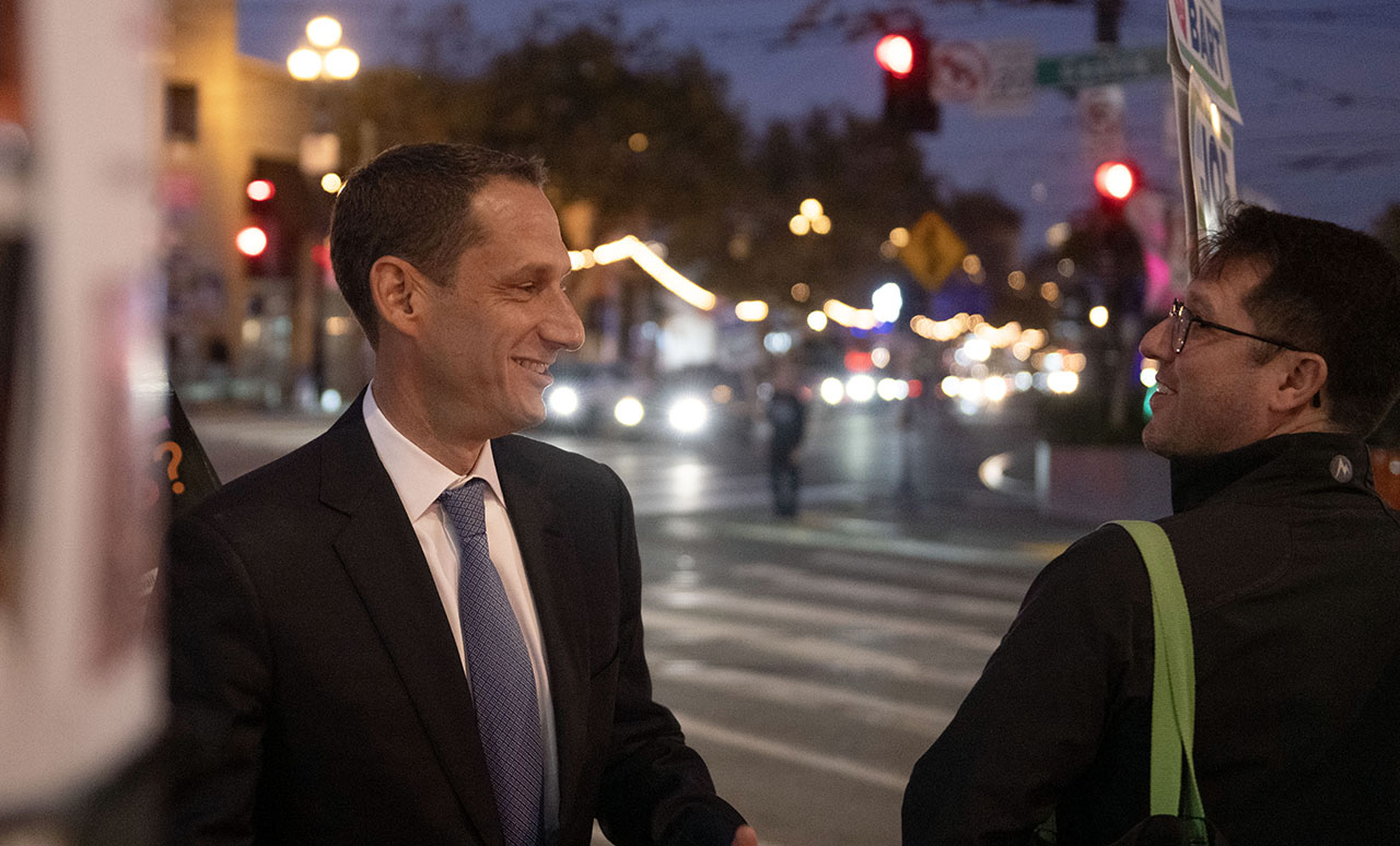 Lurie stands outside grinning as he greets a man holding a sign for BART commission candidate Joe Sangirardi