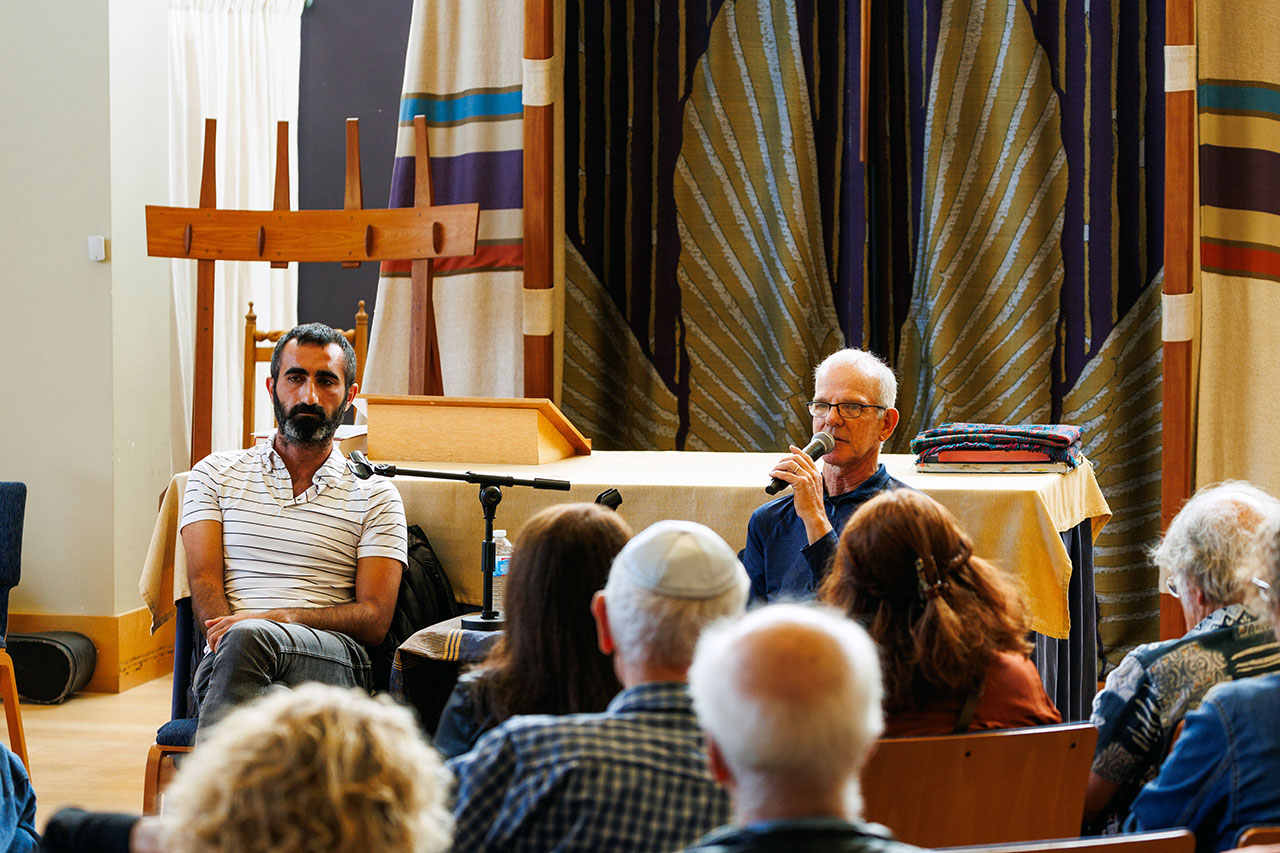 Two men sit facing a crowd. Goldberger speaks into a microphone.