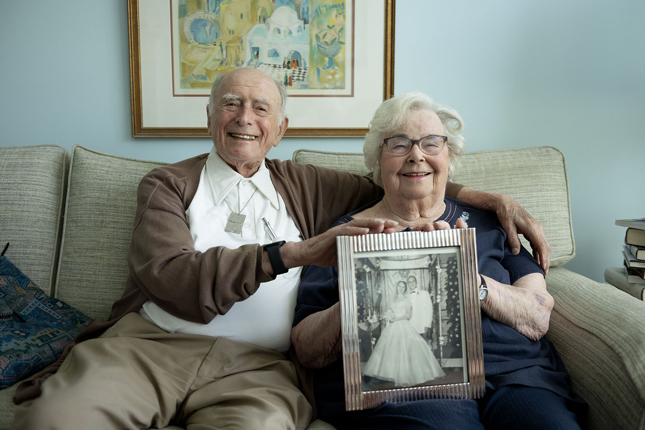 And old man and and old woman sit smiling on a couch, holding up a black and white photo of their wedding