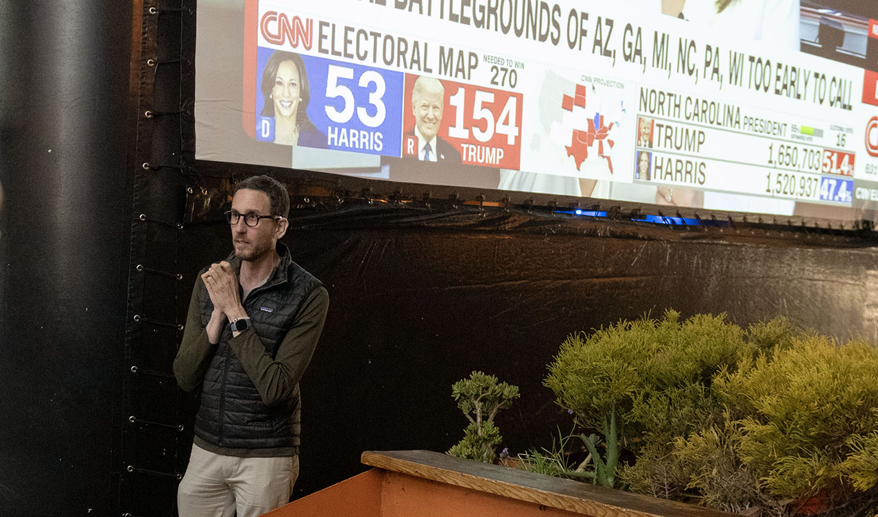 Wiener stands with his hands clasped nervously in front of a large screen showing CNN coverage of Election Day