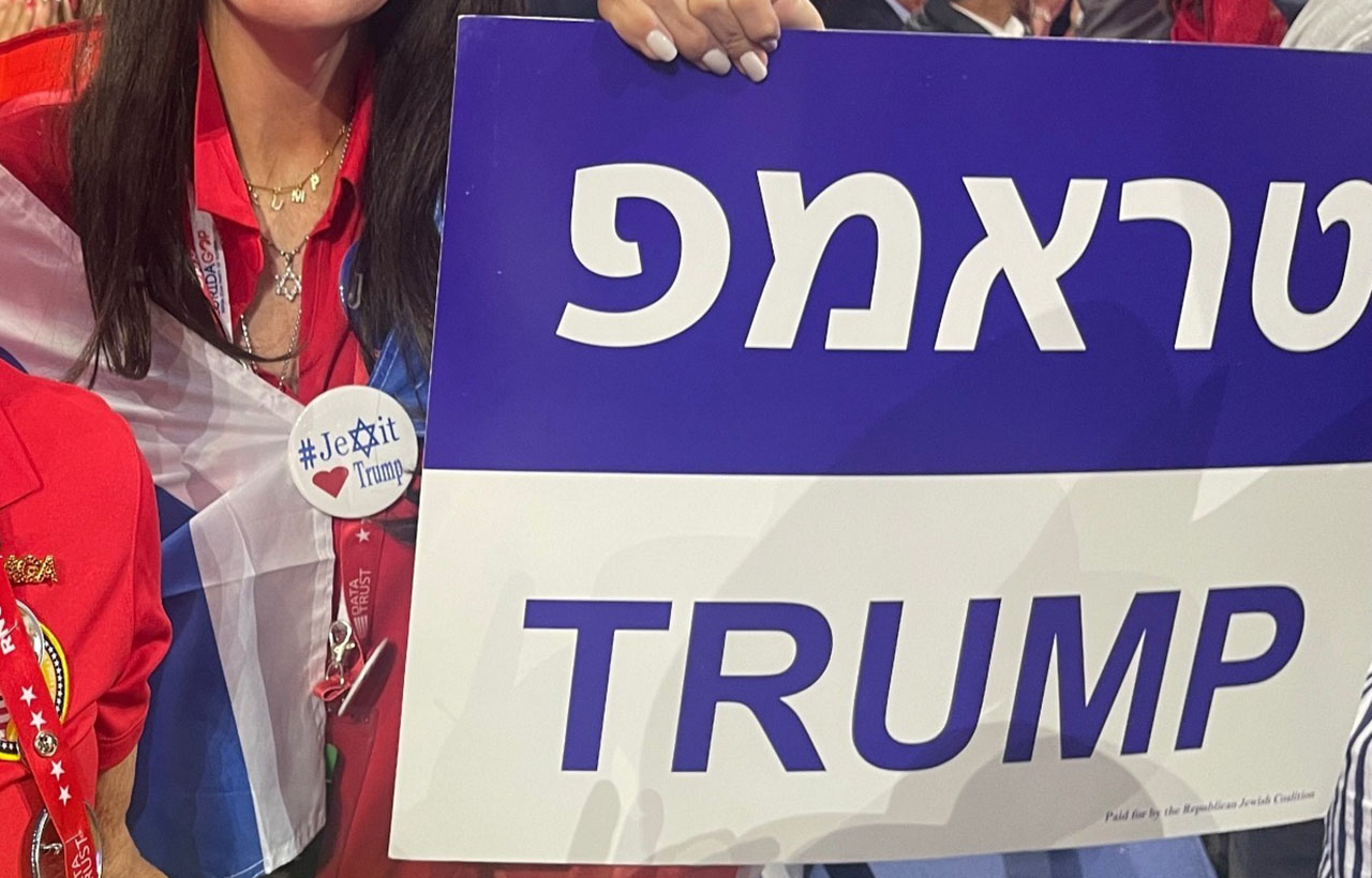 a hand holds up a sign that says "Trump" in Hebrew and English