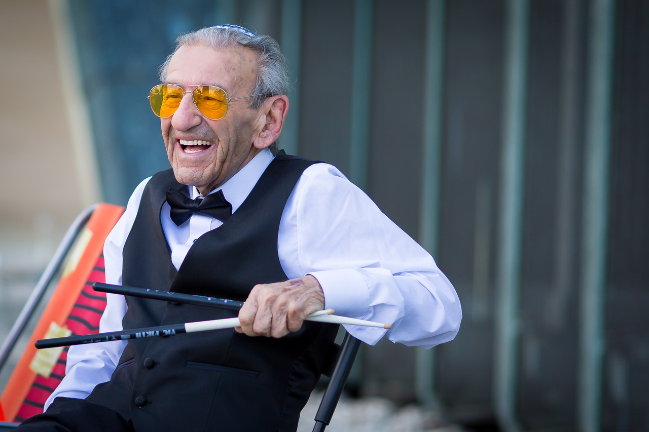 And old man sits smiling and holding a pair of drum sticks