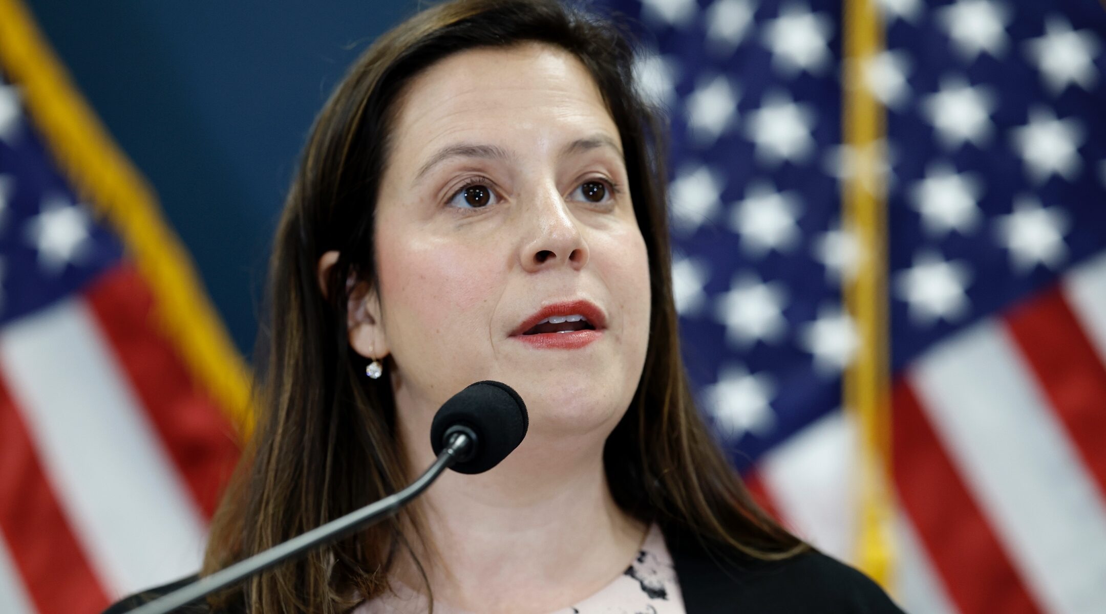 Stefanik speaks into a microphone with an American flag behind her