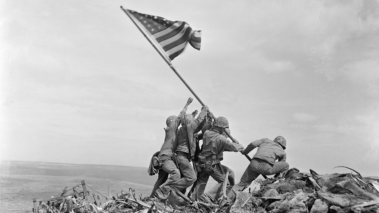 five men in WWII-era US Army combat uniforms work together to erect a flagpole atop a pile of rubble