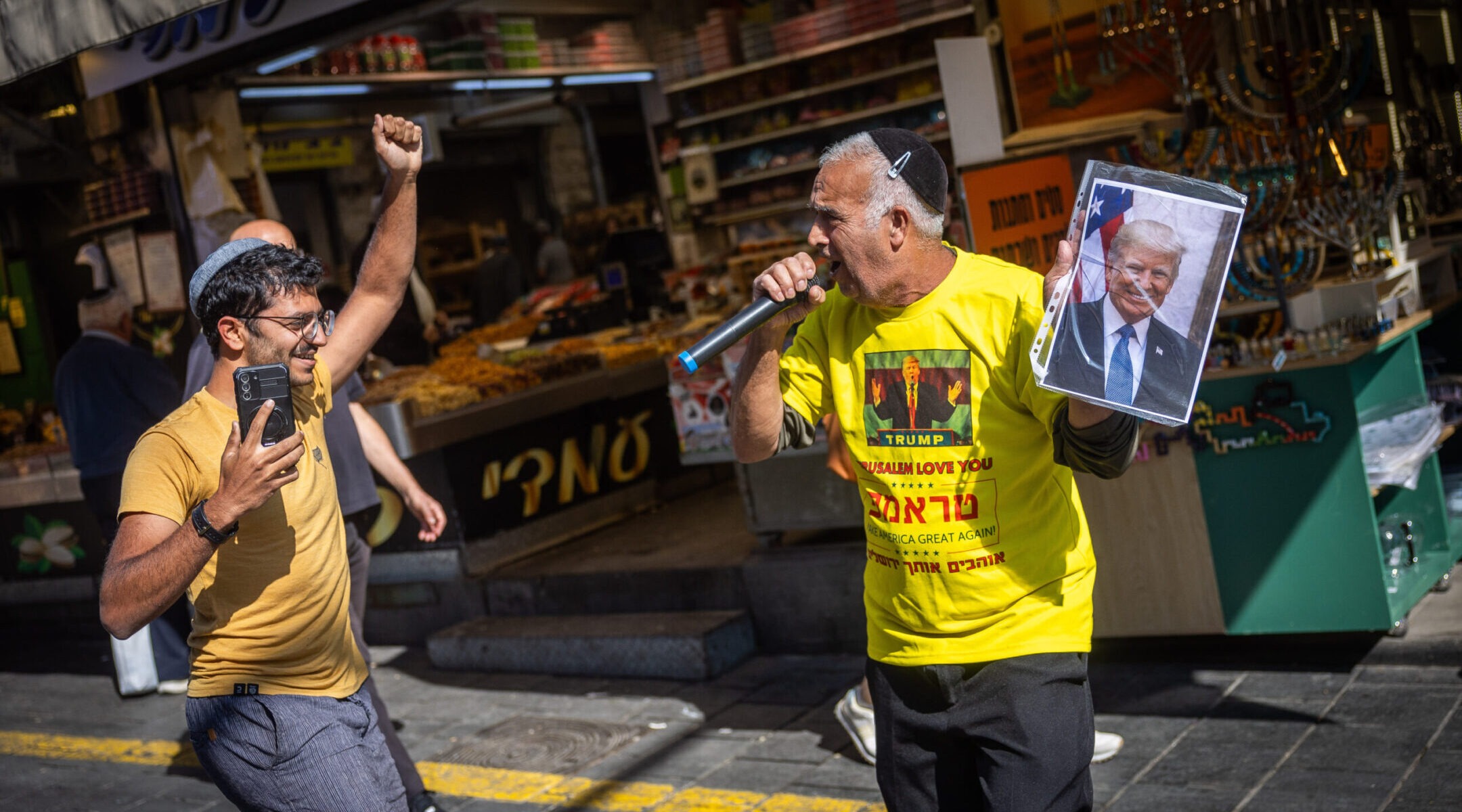 A man in a kippah and Hebrew Trump t-shirt holds up a photo of Trump and celebrates in the street with another man in a kippah