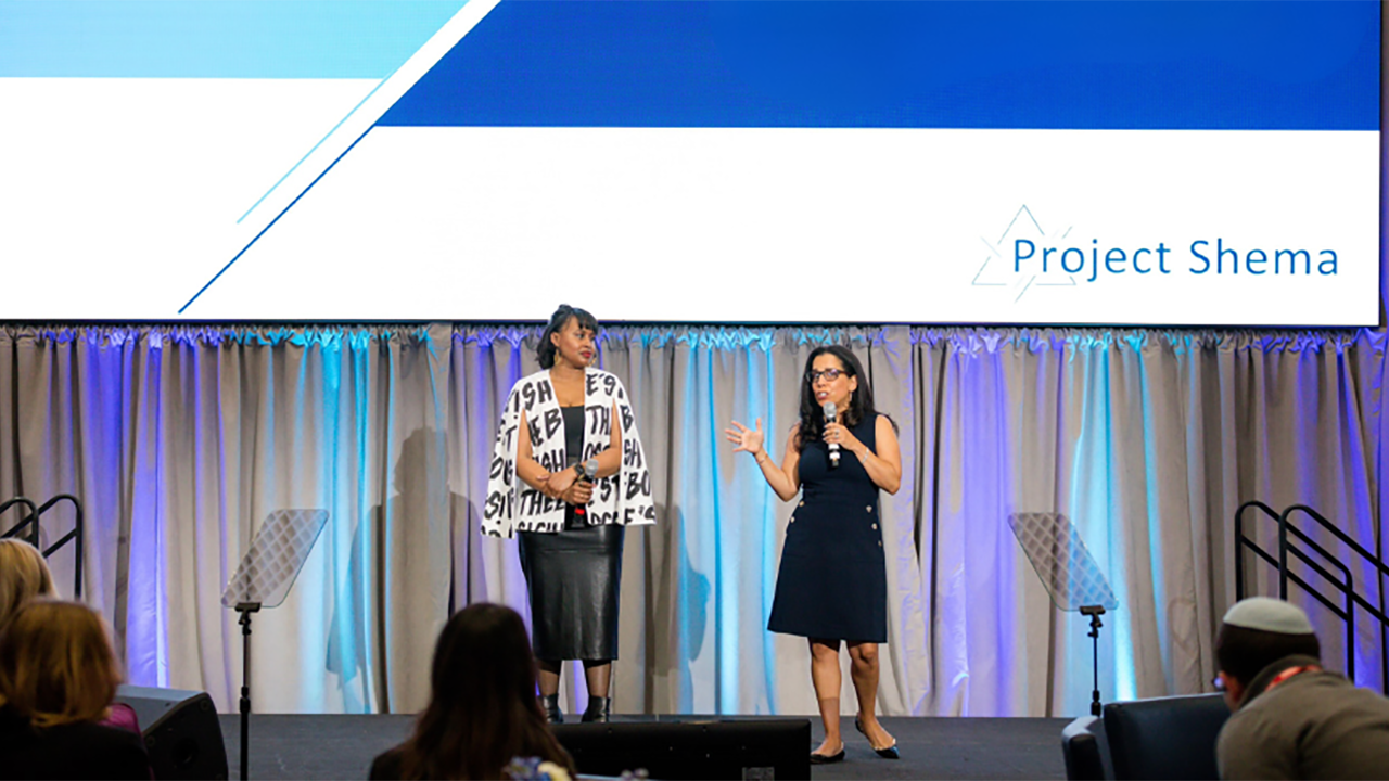 two black women stand on stage. the one on the right is speaking with a microphone. a screen behind them says "Project Shema"