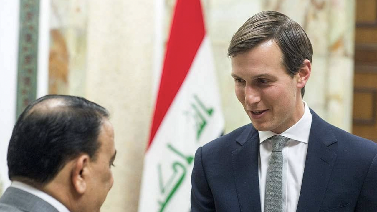 A tall white man in a suit shakes hands with a short man with brown skin. An Iraqi flag hangs in the background.