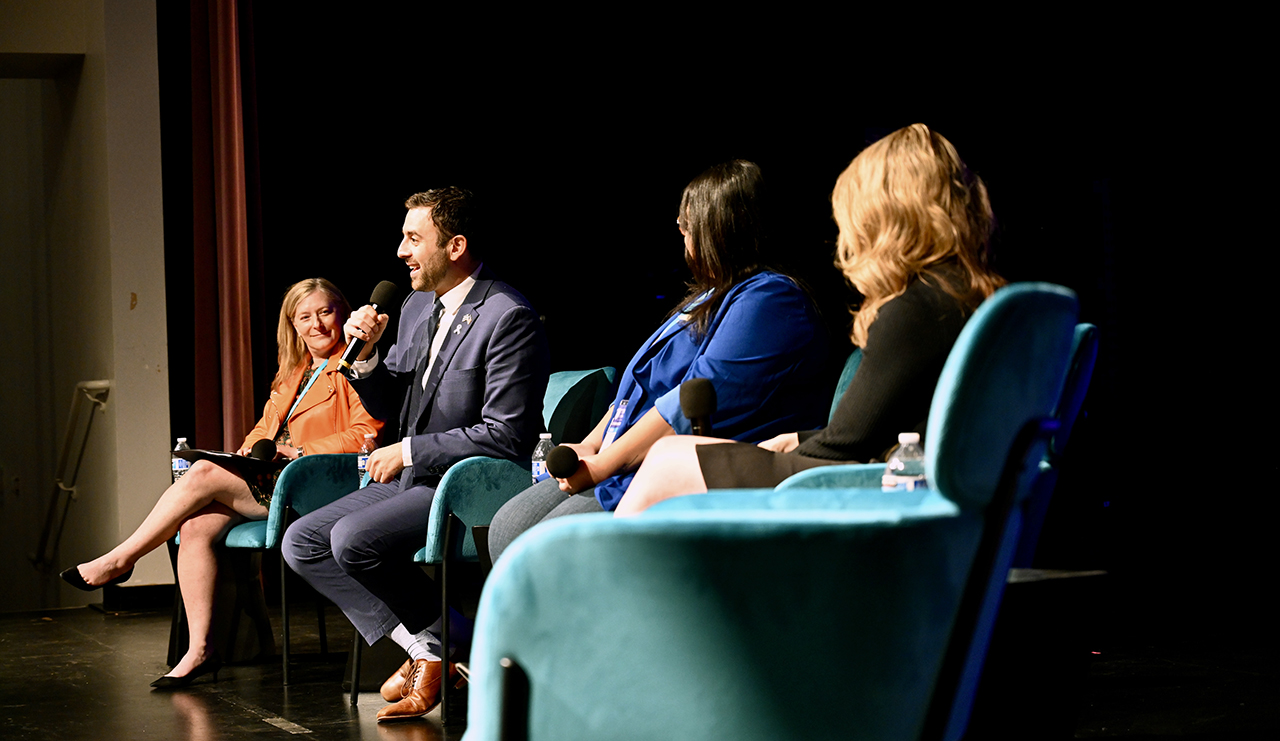 a man seated on stage speaks while flanked by three seated women who look on with interest