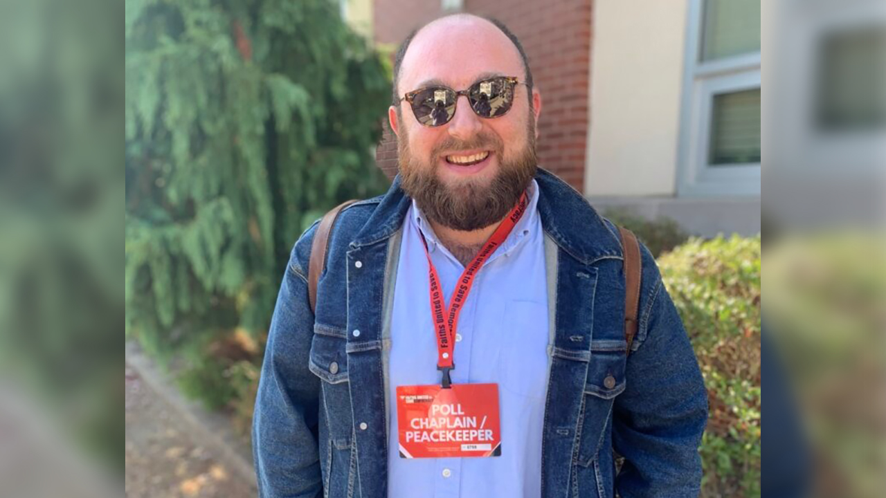 a man poses wearing a sign around his neck that says "Poll Chaplain/Peacekeeper"