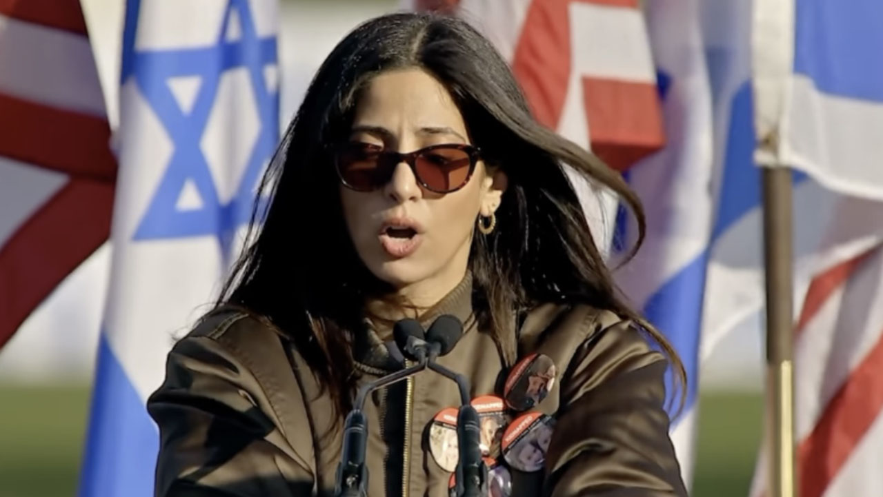 a woman in sunglasses speaks at a lectern with American and Israeli flags behind her