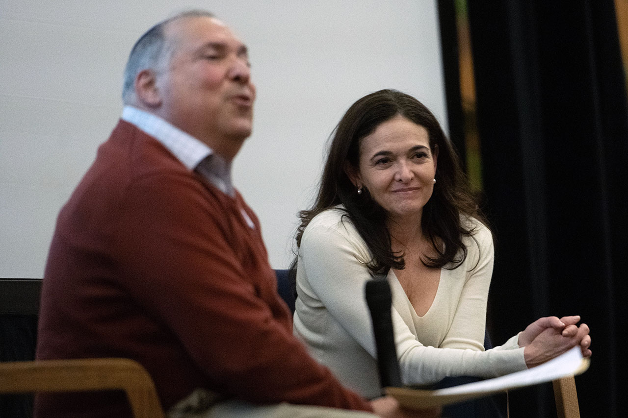 Sandberg sits next on stage next to a man who holds a microphone as her speaks
