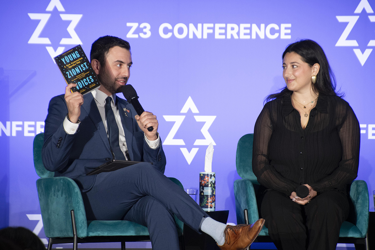 a man and a woman sit on a stage. the man is speaking into a microphone and holding up a book.