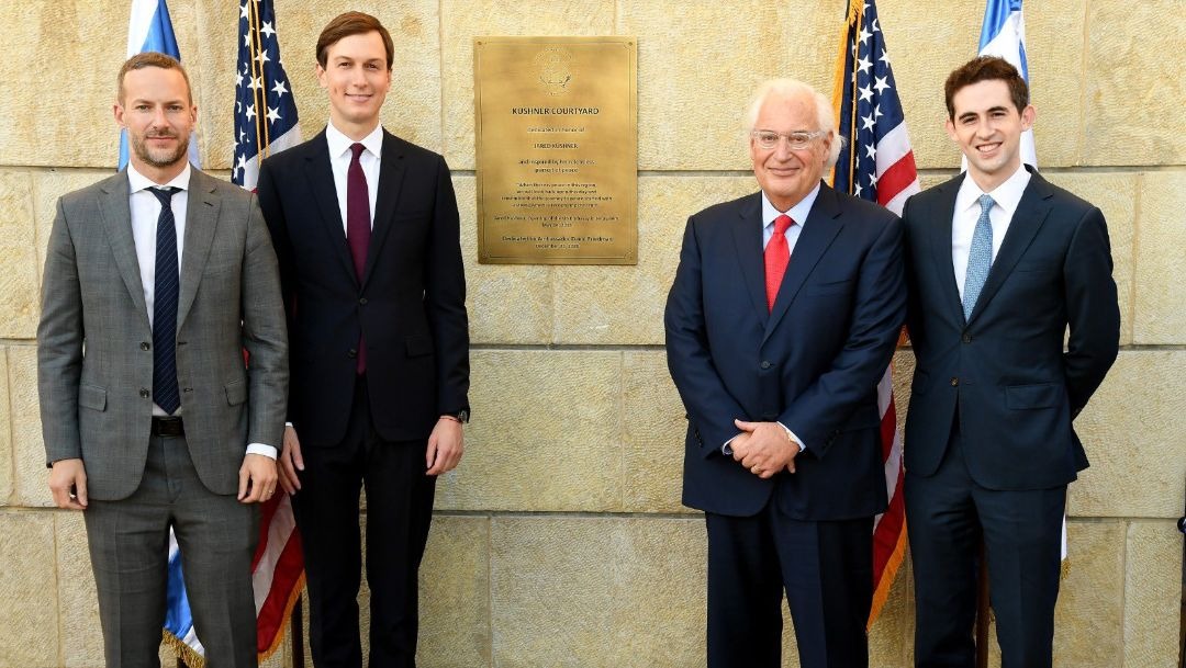 four men pose in front of a limestone wall, flanked by american flags